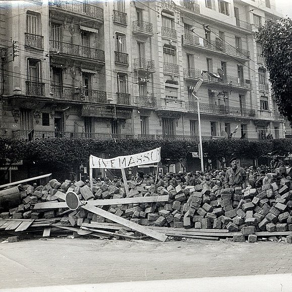L'image montre une scène historique avec un amas de briques et de débris, suggérant une barricade ou un barrissement. Au premier plan, on peut voir des personnes rassemblées, probablement des manifestants ou des membres d'une foule, observant la scène. Un panneau est visible, ce qui pourrait indiquer le message ou le motif de la manifestation. À l'arrière-plan, des bâtiments avec des balcons sont présents, typiques d'une architecture urbaine. L'atmosphère semble tendue, reflétant un moment de contestation ou de protestation.