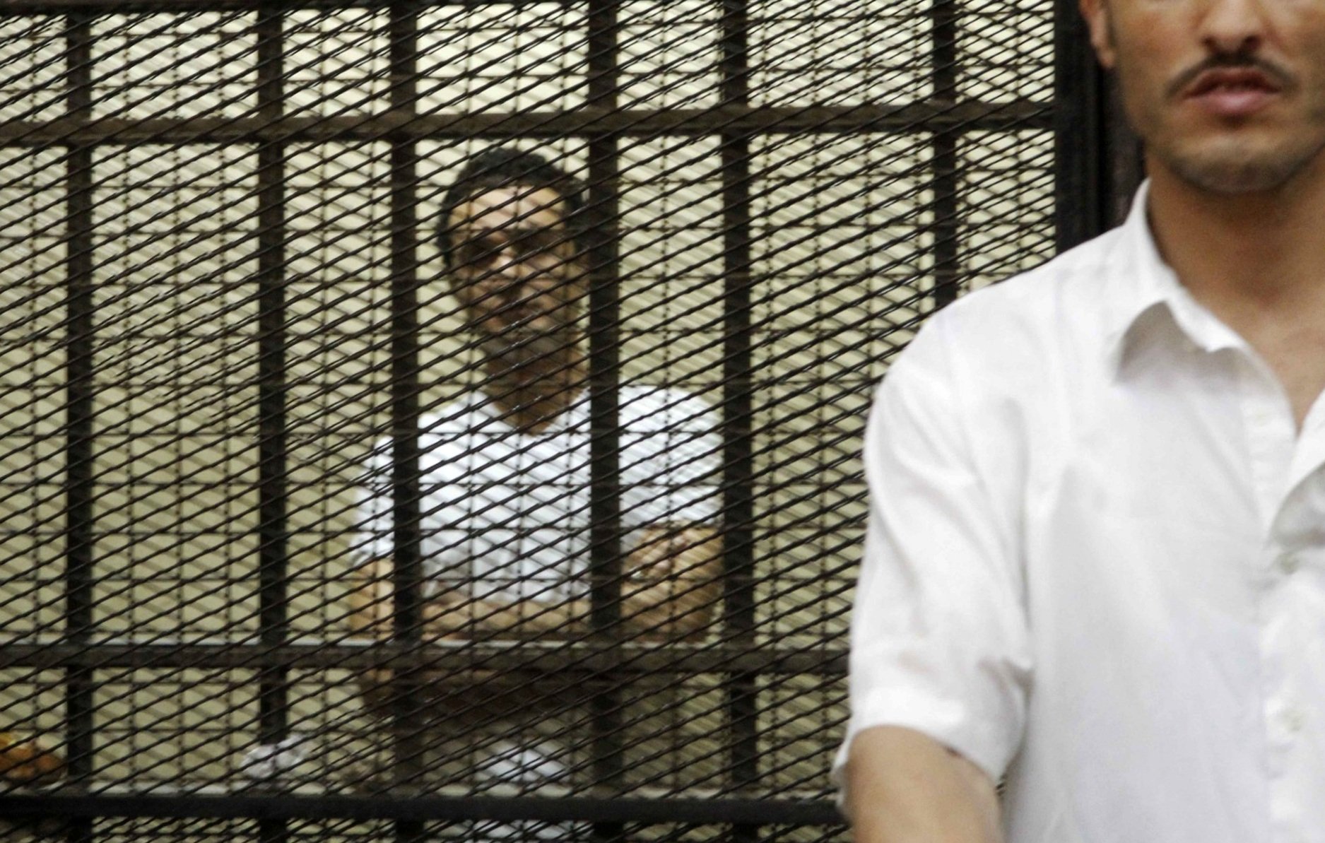 Due uomini in una stanza, uno è dietro una grata di protezione. Atmosfera tesa.