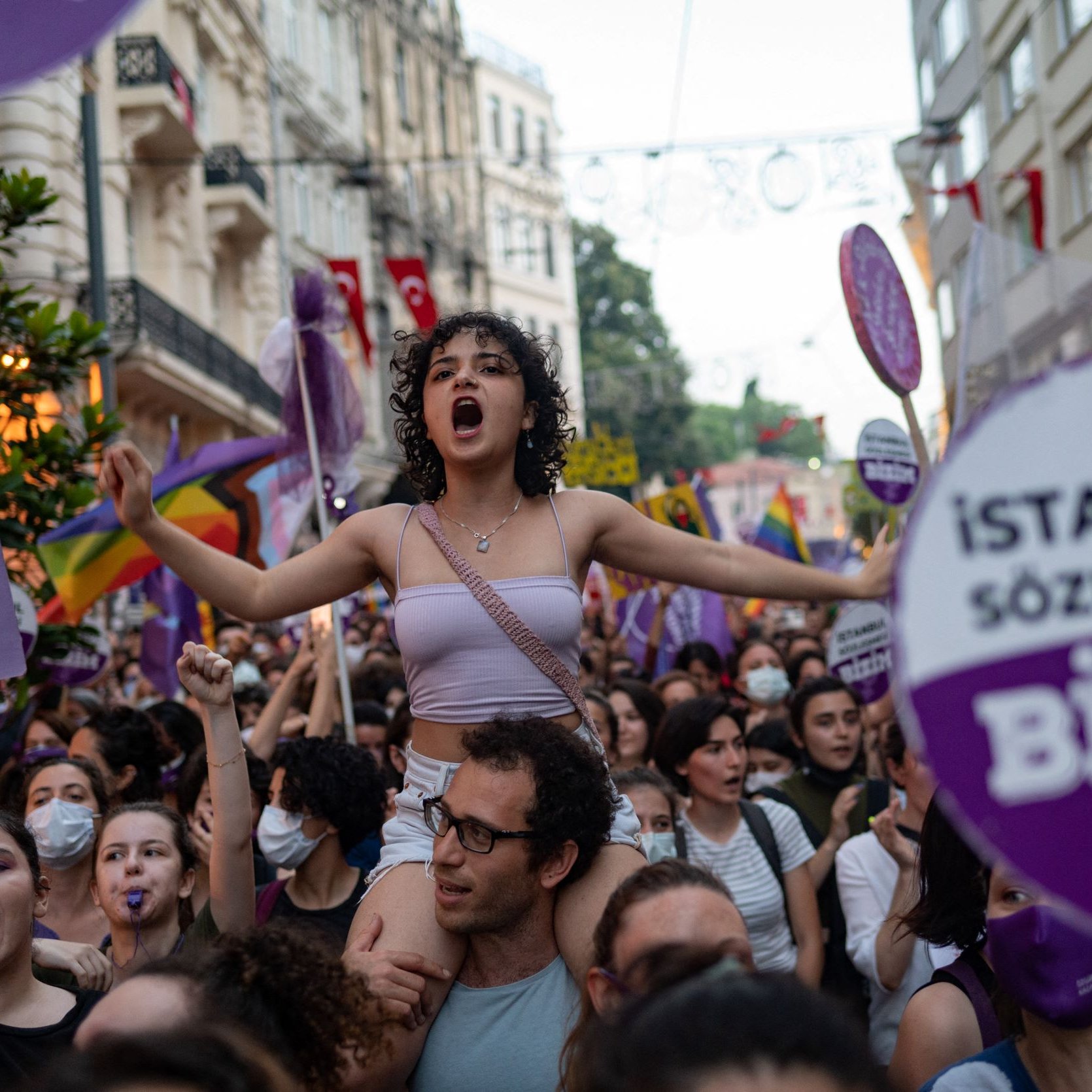 L'image montre une foule lors d'une manifestation, avec des personnes brandissant des pancartes et des drapeaux, notamment des drapeaux arc-en-ciel. Au centre, une jeune femme exprime sa détermination en chantant ou en criant, tandis qu'elle est portée par une autre personne. Tout autour, les manifestants portent des vêtements aux couleurs violettes et blanches, et l'atmosphère semble énergique et engagée. Les pancartes mentionnent des références à des droits, notamment le "contrat d'Istanbul", qui met en avant des thèmes de protection et d'égalité.