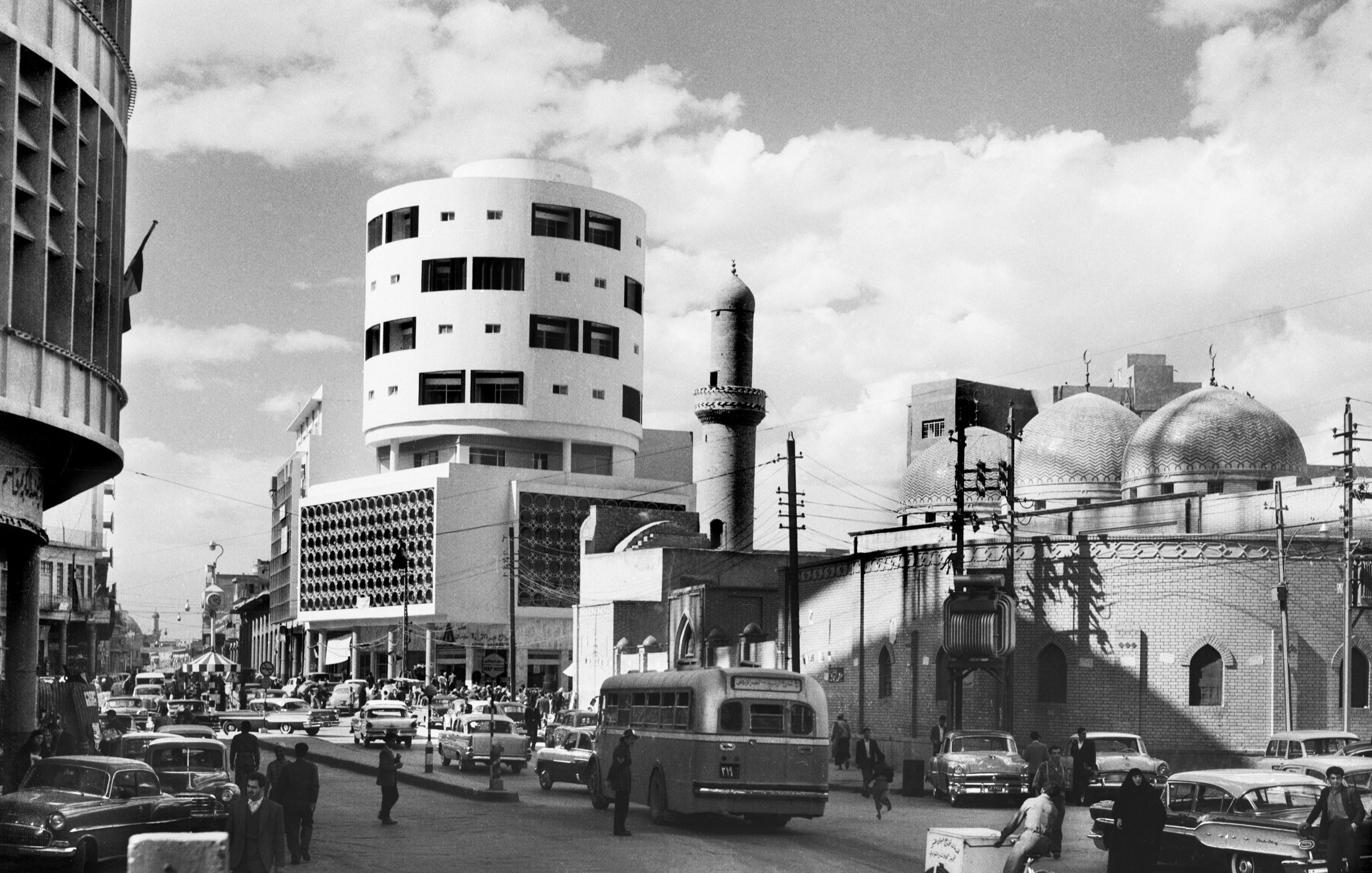 L'image en noir et blanc présente une scène urbaine animée. Au premier plan, on peut voir une rue avec plusieurs véhicules, dont des voitures et un bus. Le bâtiment à gauche a une architecture moderne, tandis qu'à droite, on aperçoit un édifice avec des dômes caractéristiques, suggérant une influence architecturale traditionnelle. Des piétons circulent, et on remarque également un homme sur un vélo. Le ciel est partiellement nuageux, ajoutant à l'atmosphère de la scène. Cette image évoque un mélange d'architecture moderne et traditionnelle dans un contexte urbain.