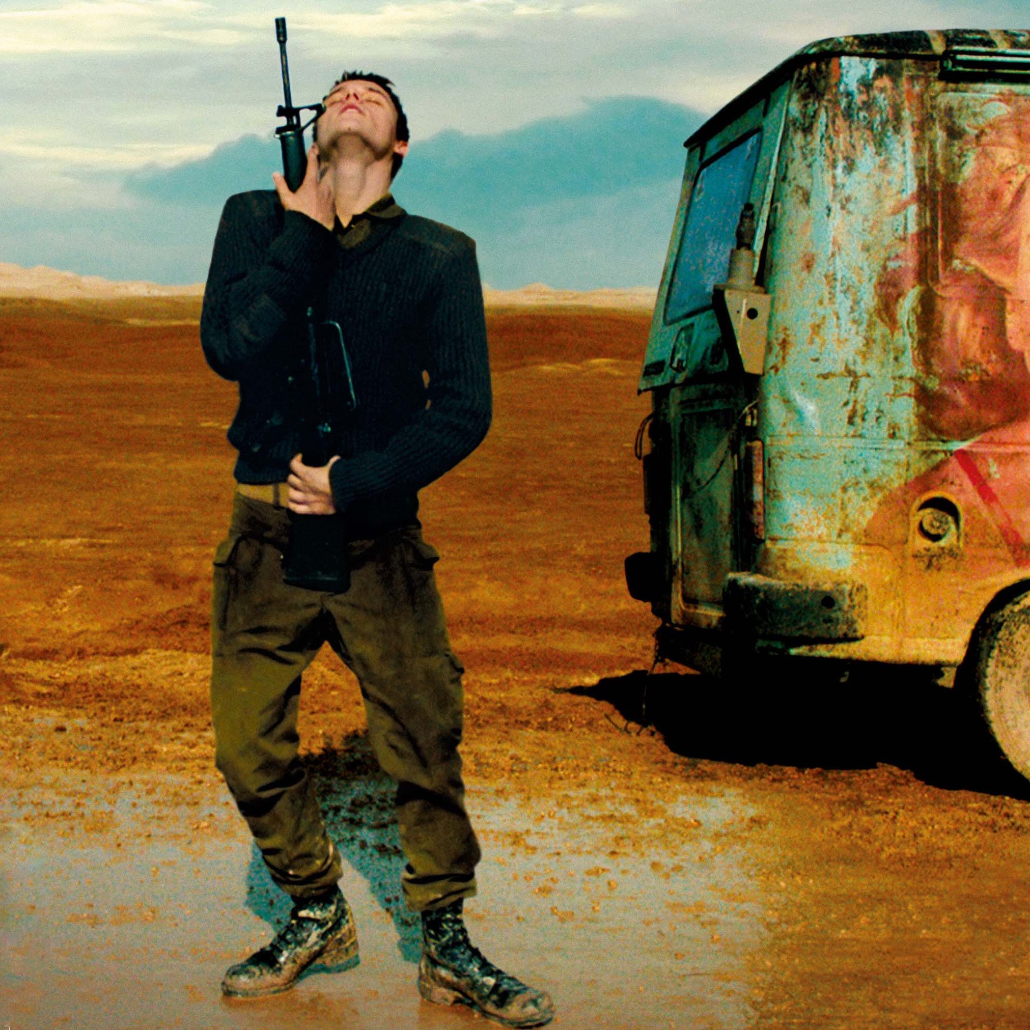 في الصورة، يظهر شاب يحمل بندقية ويرتدي ملابس عسكرية. يتblick في السماء وكأنه يعبّر عن مشاعر مختلطة من الإحباط أو الأمل. خلفه، توجد شاحنة قديمة مغطاة بالصدأ، وعلى جانبها صورة ملونة لامرأة مبتسمة تحمل مثلجات. الخلفية طبيعية، تبدو قاحلة ومملوءة بالأرض الجافة، مما يخلق تباينًا بين مشاعر الشاب وصورة الفرح على الشاحنة.