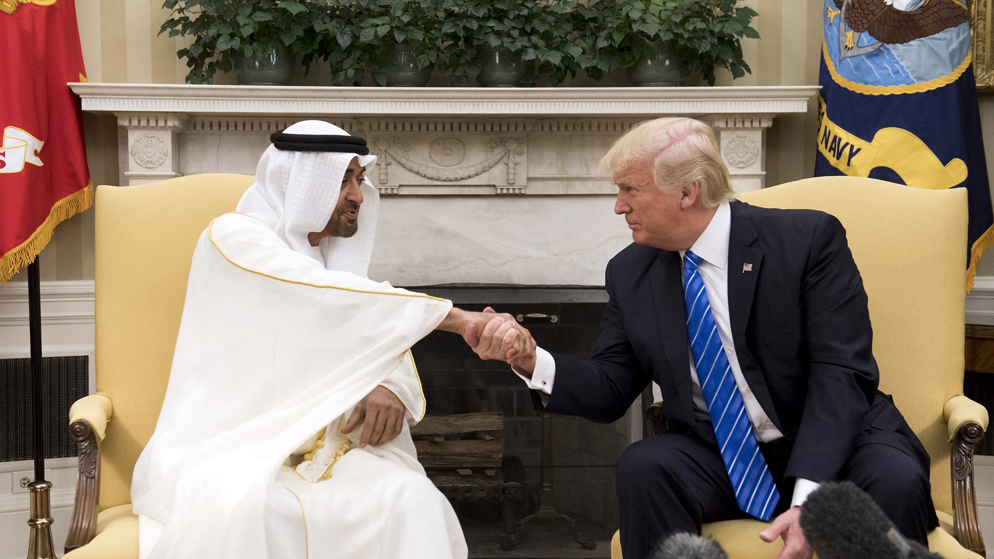 The image shows a meeting between two men, one wearing a white traditional robe and headscarf, and the other in a suit with a blue tie. They are engaged in a handshake, indicating a friendly interaction. The setting appears to be an official office, possibly the White House, with flags and decorative elements in the background. There is a focus on their handshake, showcasing a moment of diplomacy.