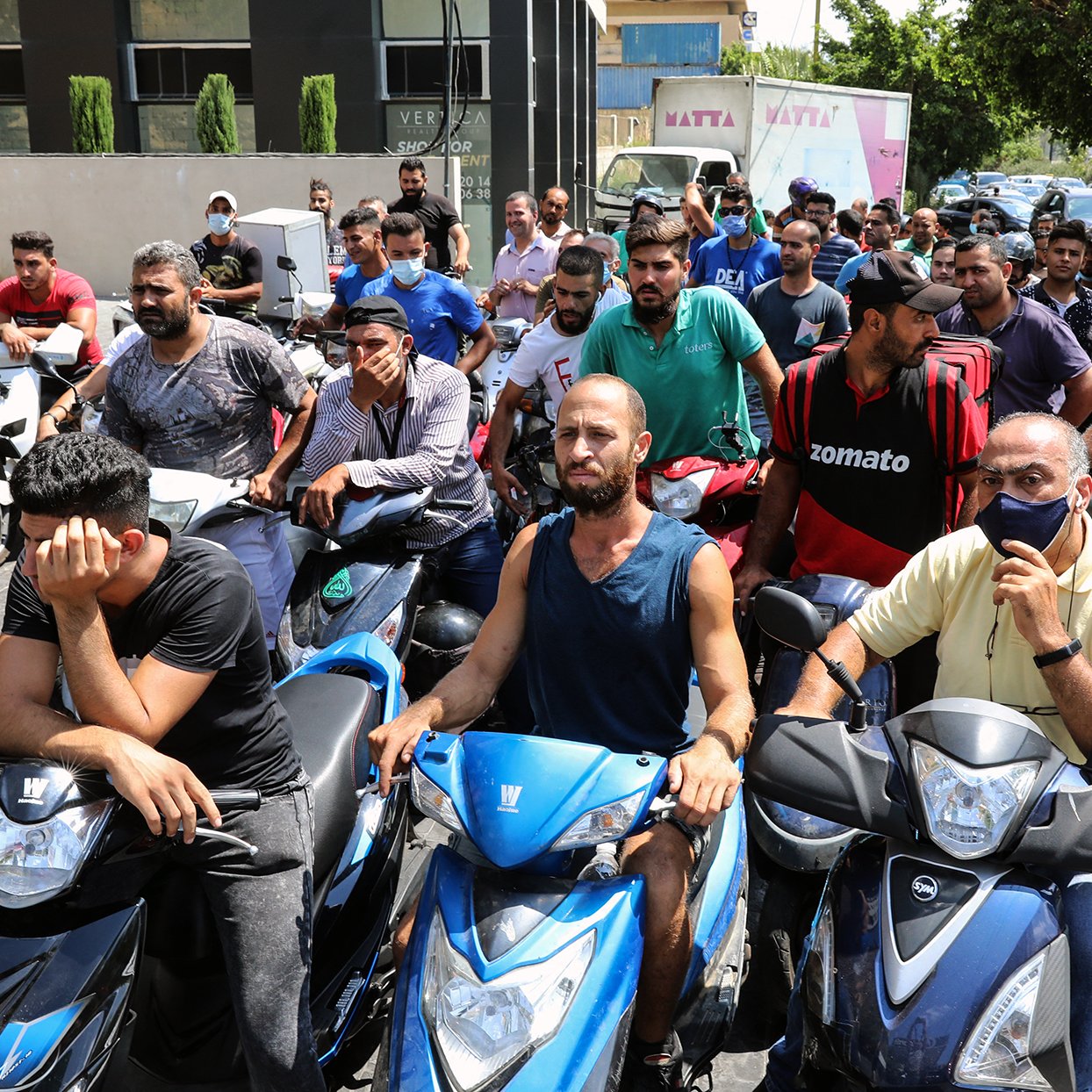 تظهر الصورة مجموعة من الأشخاص يتجمعون أمام مركز ما، حيث يبدو أنهم في انتظار الحصول على شيء ما. معظمهم يركبون دراجات نارية، ويبدو عليهم التوتر والقلق. بعضهم يظهر علامات الإحباط، بينما آخرون يتحدثون مع بعضهم. الخلفية تحتوي على مباني وأشجار، مما يشير إلى أن المشهد في منطقة حضرية.