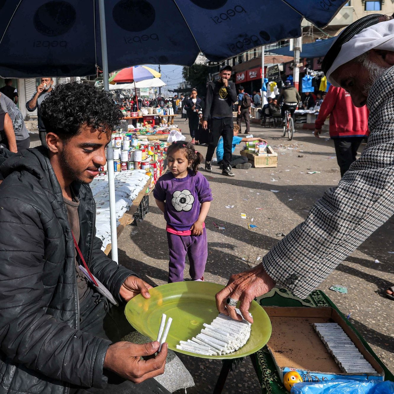 L'image montre une scène de marché dans une rue animée. Un jeune homme est assis sur un banc, tenant une assiette ronde verte sur ses genoux, où semblent être disposés des articles, probablement des bonbons ou des bâtonnets. Un homme plus âgé s'approche de lui, tenant quelque chose dans sa main, peut-être en train de lui faire une offre ou de lui donner des articles. En arrière-plan, il y a des gens qui se déplacent, ainsi que des étals avec divers produits. Une petite fille se tient à proximité, observant la scène. Les parasols créent une ambiance de marché, tandis que l'atmosphère semble vivante et animée.