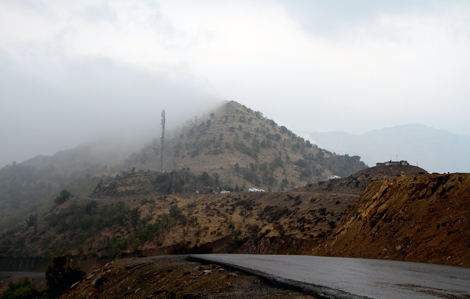 تظهر في الصورة مشهدًا جبليًا يكتنفه الضباب، حيث تتساقط السحب بشكل كثيف على الجبل. في الجزء العلوي من الجبل، يمكن رؤية برج اتصالات. الطريق يلتف حول الجبل، ويظهر بشكل مائل مع اختلافات في الألوان بين الصخور والتربة. الأجواء تبدو هادئة، مع لمسات من الغموض بسبب الضباب.