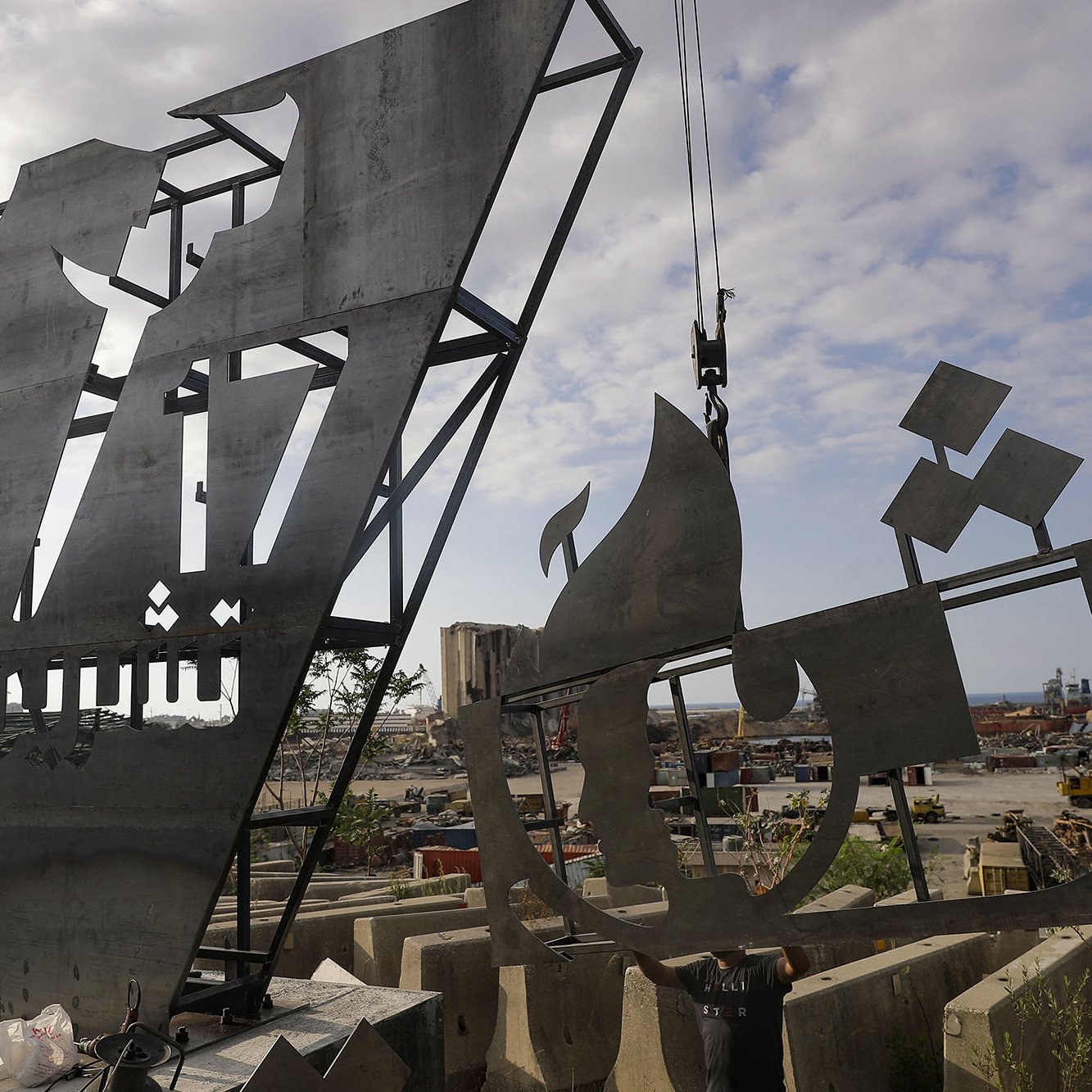 The image depicts two large, metallic sculptures or structures under construction or installation in an industrial outdoor setting. The sculptures show abstract forms, one resembling a stylized letter or shape and the other possibly depicting a ship or vessel. The background features a harbor with cranes and construction materials scattered around, suggesting an active worksite. The sky is partly cloudy, adding to the overall atmosphere of a busy environment.