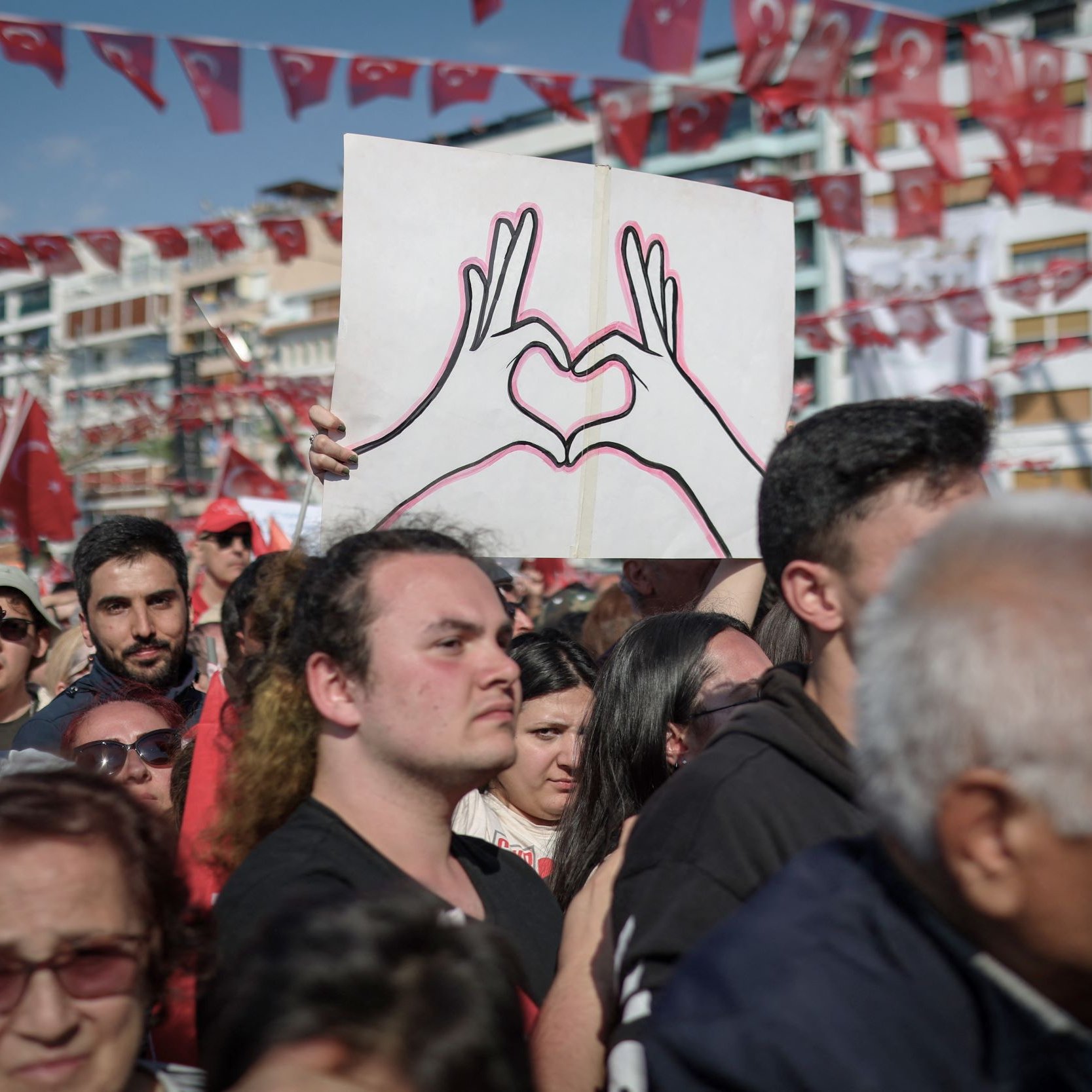 L'image montre une grande foule rassemblée lors d'un événement en plein air. Au centre, une personne tient une pancarte avec un dessin de deux mains formant un cœur. La majorité des personnes présentes portent des vêtements aux couleurs nationales, notamment du rouge et du blanc, et certains affichent des drapeaux turcs. L'ambiance semble festive et engagée, témoignant d'une manifestation ou d'une célébration collective.