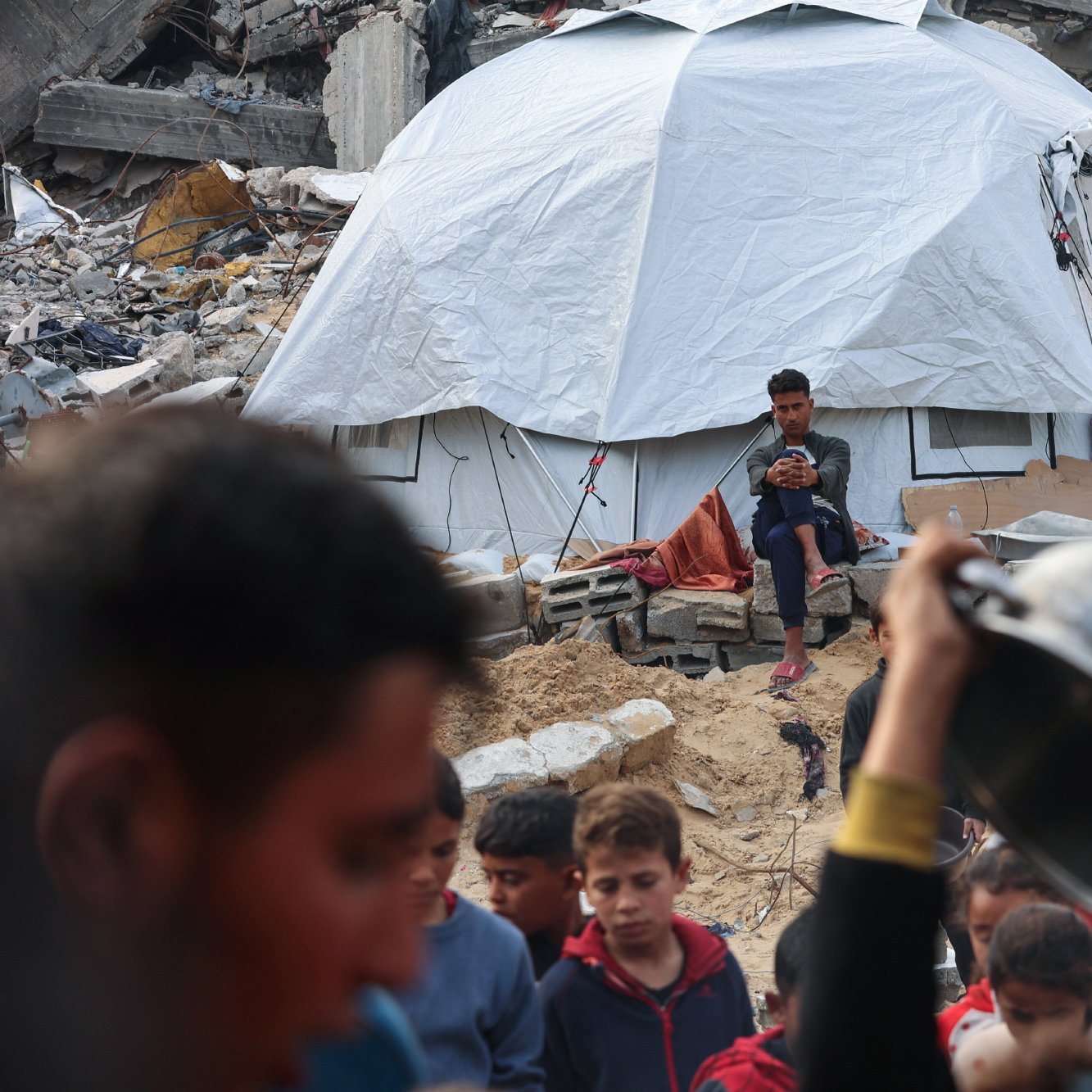 در این تصویر، یک چادر سفید در میان خرابه‌ها قرار دارد و یک نوجوان در حال نشستن و استفاده از گوشی تلفن همراه است. در جلوی چادر، یک گروه از کودکان و افراد بزرگسال دیده می‌شوند که به سمت دوربین نگاه می‌کنند یا در حال انجام کارهایی هستند. زباله‌ها و خرابه‌های ساختمان‌های تخریب‌شده در پس‌زمینه به وضوح احساس ناامیدی و بحران را منتقل می‌کنند.