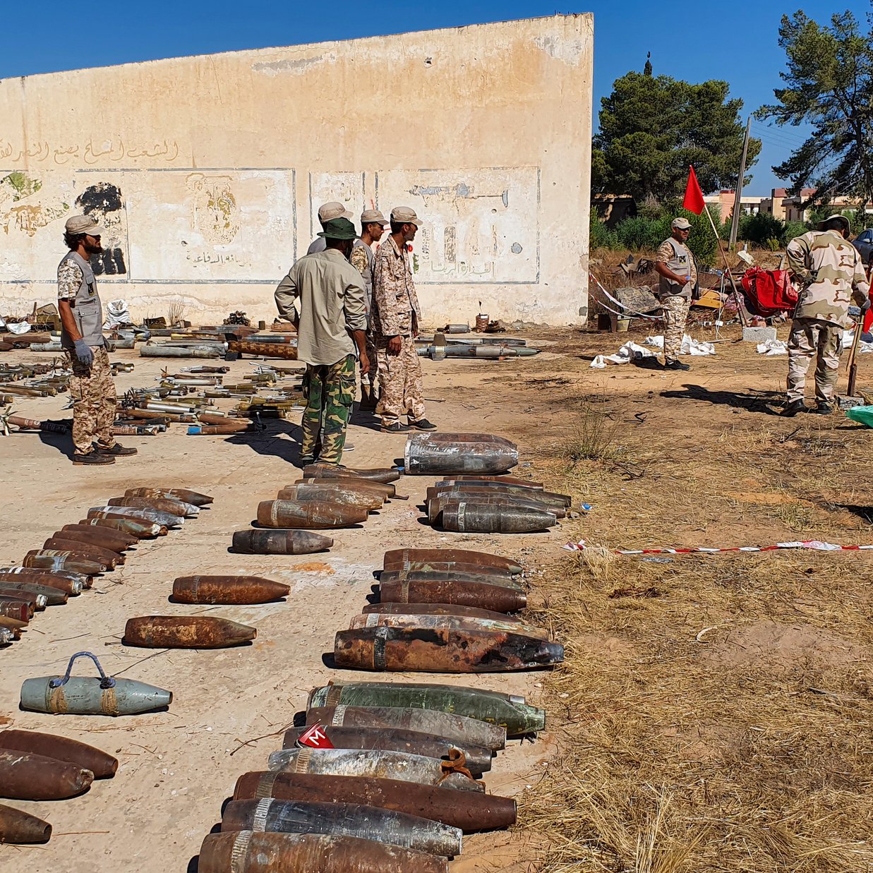 L'image montre un site en extérieur où des bombes et des munitions sont disposées au sol. Plusieurs personnes en uniformes militaires sont présentes, certaines examinant les objets, tandis que d'autres semblent discuter ou travailler sur le terrain. En arrière-plan, on peut voir un mur partiellement recouvert de graffitis et des arbres qui entourent la zone. Des drapeaux sont également visibles, et le sol est marqué par des restes de munitions, signifiant probablement un site de déminage ou de collecte de matériel explosif.