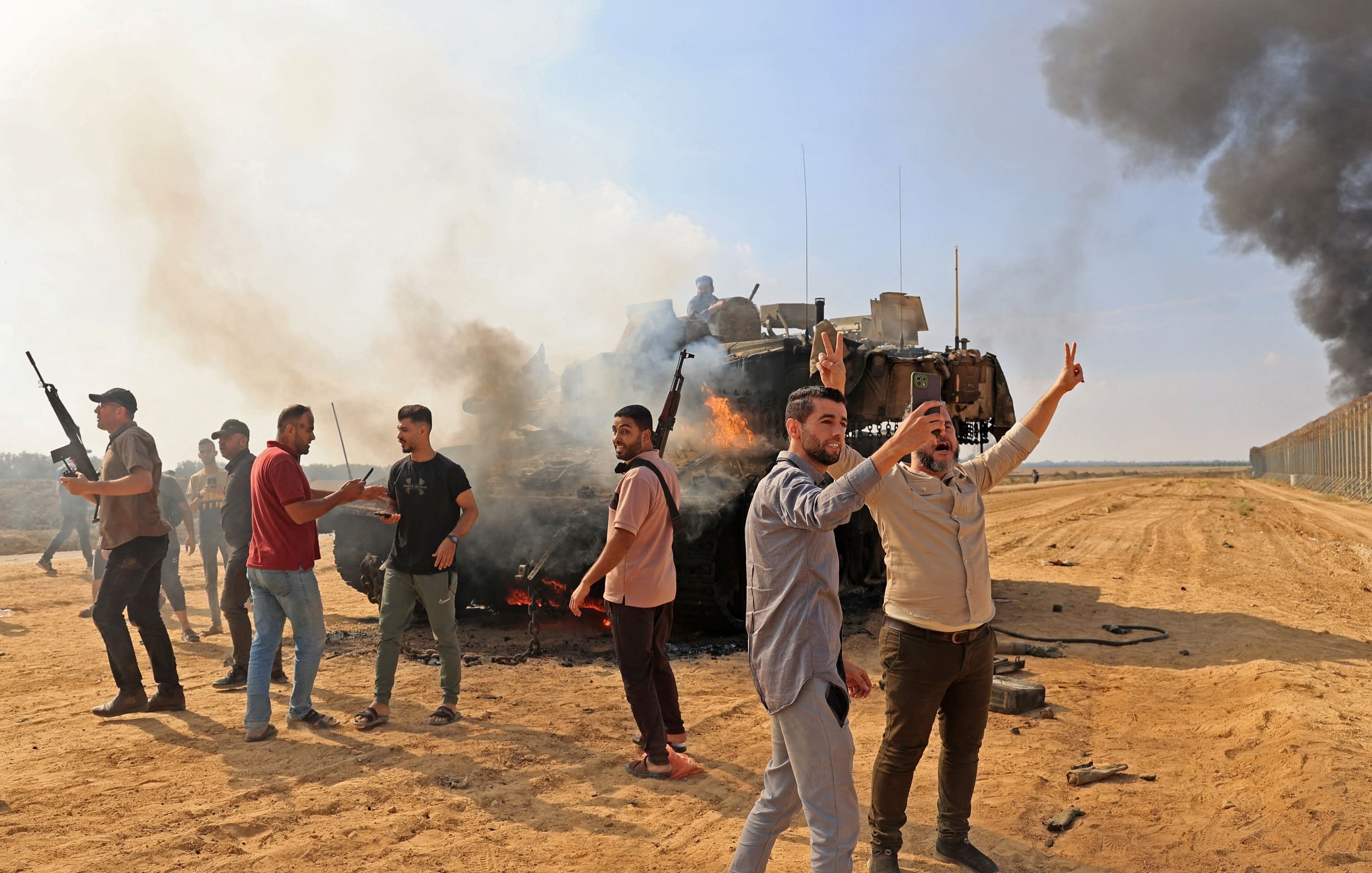 در تصویر، تعدادی از افراد در یک منطقه بیابانی مشاهده می‌شوند که به سمت دوربین اشاره می‌کنند و برخی از آن‌ها سلاح به‌دست دارند. در پس‌زمینه، دود سیاه و غلیظی از یک وسیله نقلیه نظامی در حال سوختن دیده می‌شود. به نظر می‌رسد که این صحنه به یک وقایع پرتنش یا بحران های اجتماعی مربوط می‌شود. افراد در حال ابراز هیجان و یا اعتراض هستند و فضای کلی تصویر، حالتی داغ و ناآرام دارد.