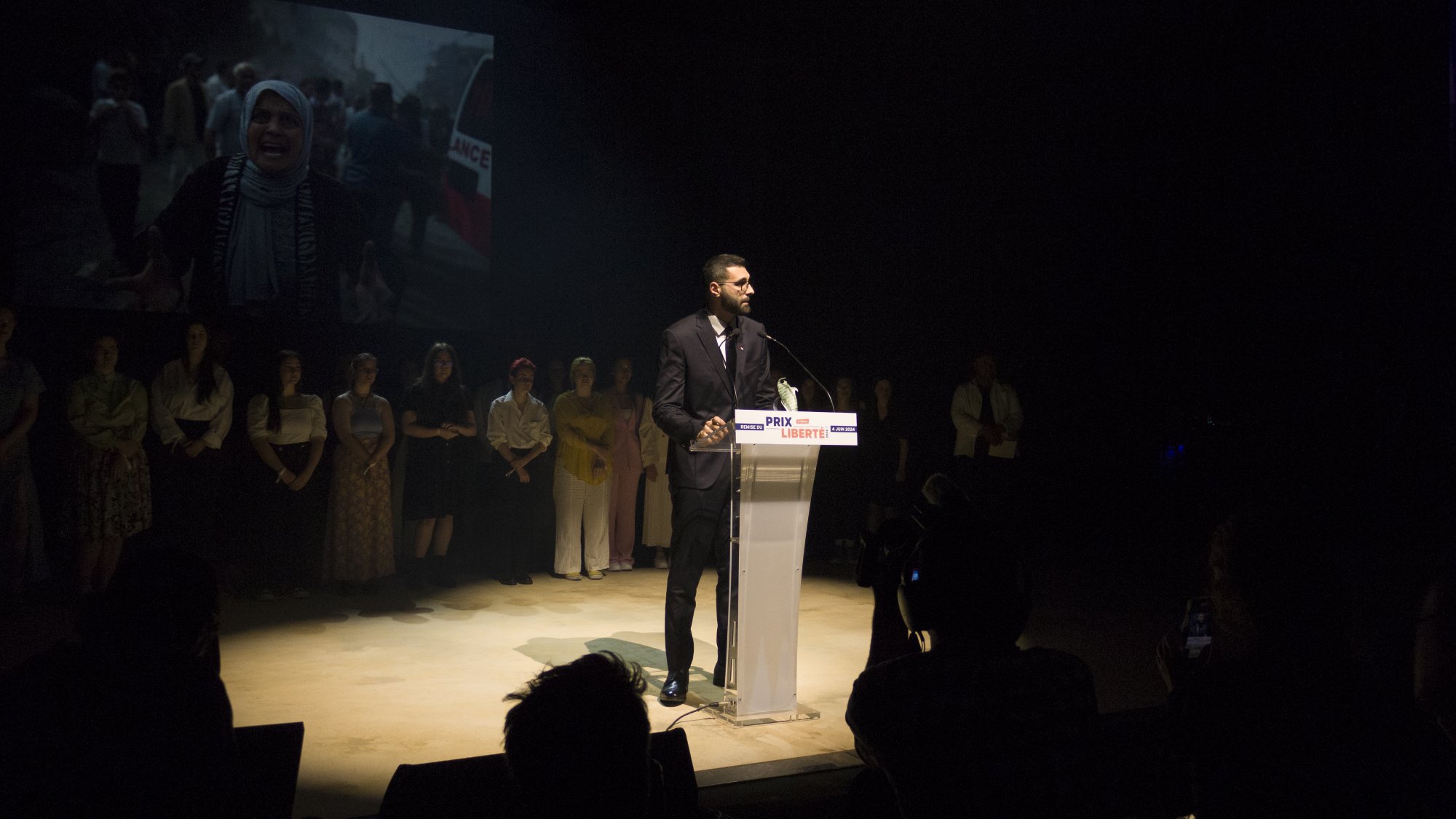 L'image montre un homme debout à un pupitre transparent, prenant la parole lors d'un événement. Il est habillé en costume et semble s'adresser à un public. En arrière-plan, on peut voir un écran projetant une vidéo ou une image, vraisemblablement liée à un sujet important, étant donné l'expression des personnes présentes. Au premier plan, on peut apercevoir des membres du public et des photographes, créant une atmosphère de concentration et d'engagement. L'éclairage met en évidence l'orateur, tandis que le reste de la scène est plus sombre.