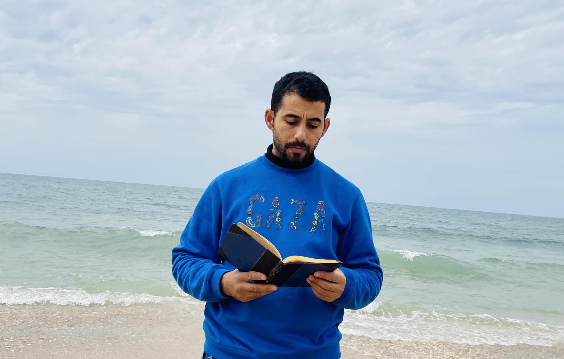 L'image montre un homme debout sur une plage, face à la mer. Il porte un pull bleu avec "GAZA" écrit dessus. Il tient un livre ouvert dans ses mains et semble lire ou se concentrer sur son texte. En arrière-plan, on peut voir des vagues qui se brisent doucement sur le rivage, et le ciel est nuageux, ajoutant une atmosphère calme à la scène.