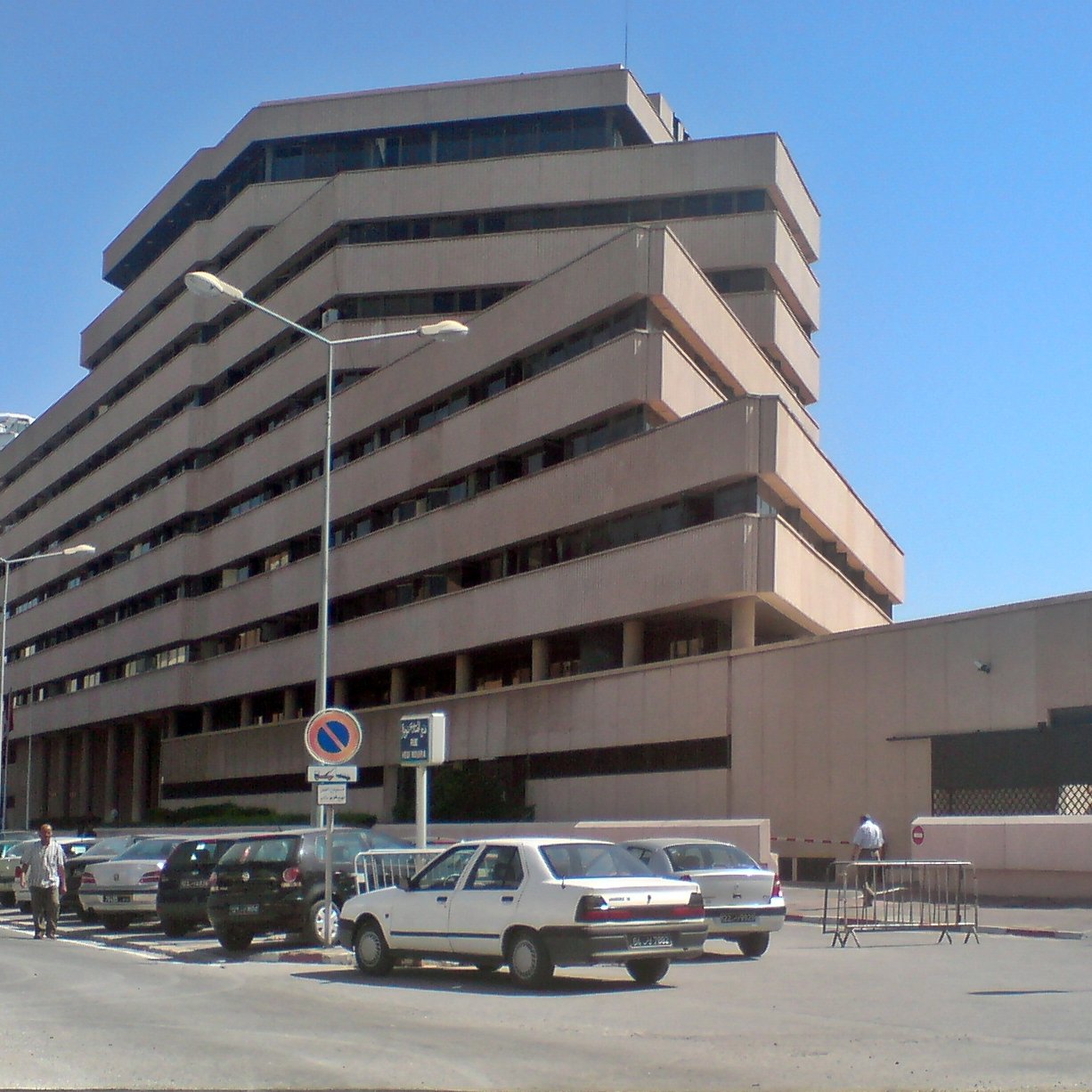 L'image montre un bâtiment moderne à plusieurs étages avec une architecture angulaire et des balcons en saillie. La façade est en béton, avec des couleurs neutres. On peut voir des voitures garées le long de la route, et il semble y avoir des piétons dans la zone. Le ciel est dégagé, ce qui donne une atmosphère ensoleillée à la scène. Le bâtiment dégage une impression de fonctionnalité et de modernité.