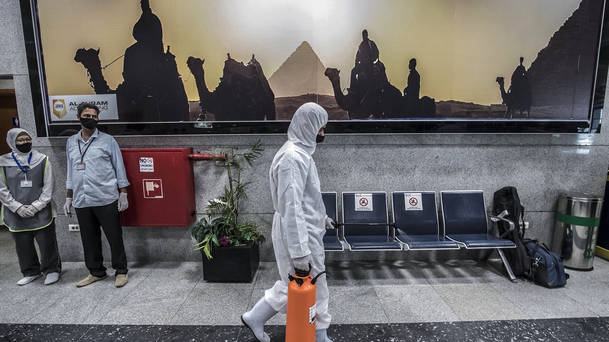 L'image montre un hall d'aéroport ou une station, avec un fond représentant des silhouettes de chameaux et de pyramides, visiblement au coucher du soleil. Au premier plan, une personne en costume de protection blanc marche, tenant une valise orange. Deux personnes en uniforme, probablement du personnel de sécurité ou de santé, se tiennent sur le côté, portant des masques. L'environnement est moderne et propre, avec des sièges en métal et des éléments de décoration comme des plantes.