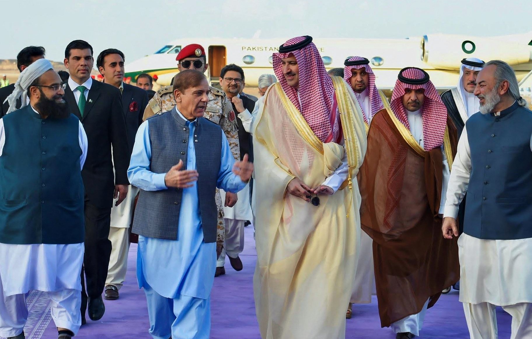 En la imagen, se puede ver a un grupo de hombres en un evento formal. Uno de ellos, vestido con una túnica azul, parece estar conversando animadamente mientras camina. Los demás, vestidos con trajes tradicionales y de gala, incluyen a personas con vestimenta árabe y otros trajes formales. Están rodeados de un ambiente elegante, con un jet privado y una alfombra morada en el fondo. La escena sugiere una ocasión diplomática o una visita de estado.