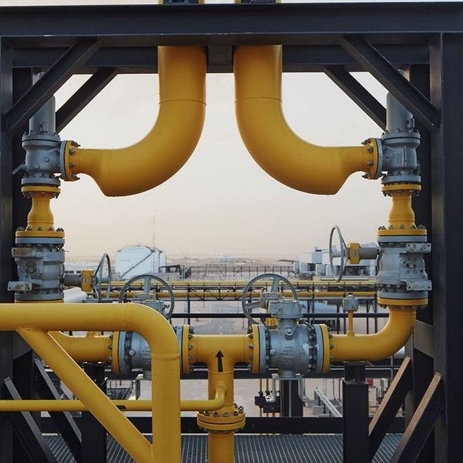 L'image montre un réseau de tuyauterie industrielle. On peut voir des tuyaux en métal, peints en jaune, qui se croisent et se connectent à différentes valves et équipements. L'arrière-plan présente un paysage industriel, avec d'autres structures et installations visibles au loin, suggérant un environnement de production ou de traitement de ressources. Le tout semble bien organisé et conçu pour la circulation de fluides, probablement dans le secteur pétrolier ou gazier.