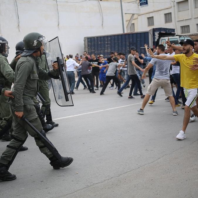 L'image montre une scène de confrontation entre des agents de sécurité en uniforme, portant des casques et des protections, et un groupe de manifestants. Les policiers avancent avec des boucliers, tandis que des personnes dans le public semblent manifester avec des gestes de défi. L'atmosphère est tendue, et le cadre urbain suggère un rassemblement intense, possiblement dans un contexte de protestation ou de manifestation. Les participants sont habillés de manière casual, et on peut voir une diversité dans leurs attitudes et leurs réactions face à la situation.