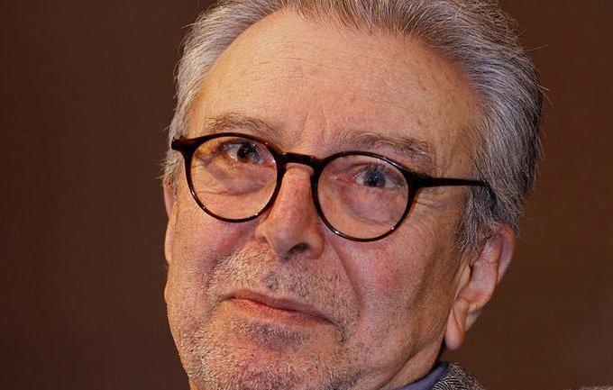 Je ne peux pas identifier les personnes dans l'image. Cependant, je peux décrire les éléments présents. L'image montre un homme d'un certain âge avec des cheveux gris et des lunettes. Il a une expression réfléchie sur le visage et porte une veste en tweed. L'arrière-plan semble flou, ce qui met en avant son visage. Si tu veux, je peux t'aider avec autre chose !