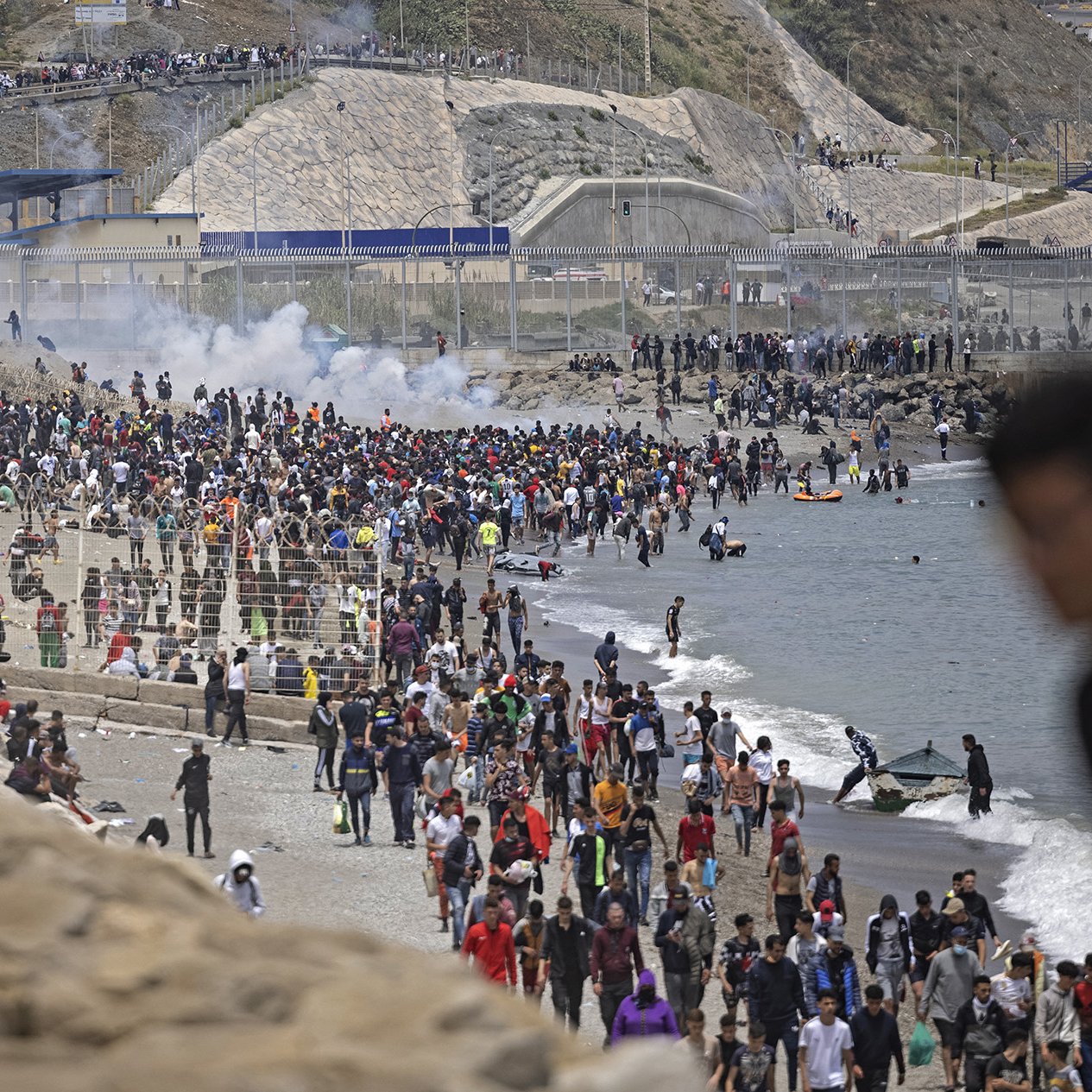 L'image montre une foule nombreuse sur une plage, avec des personnes rassemblées près de l'eau et d'autres sur le rivage. En arrière-plan, on peut voir des collines ainsi que des structures de béton. Il y a également des éléments de chaos, comme de la fumée, ce qui pourrait indiquer une situation tendue ou un événement significatif. Des gens semblent se déplacer, certains en direction de la mer, tandis que d'autres se trouvent sur le sable ou dans l'eau.