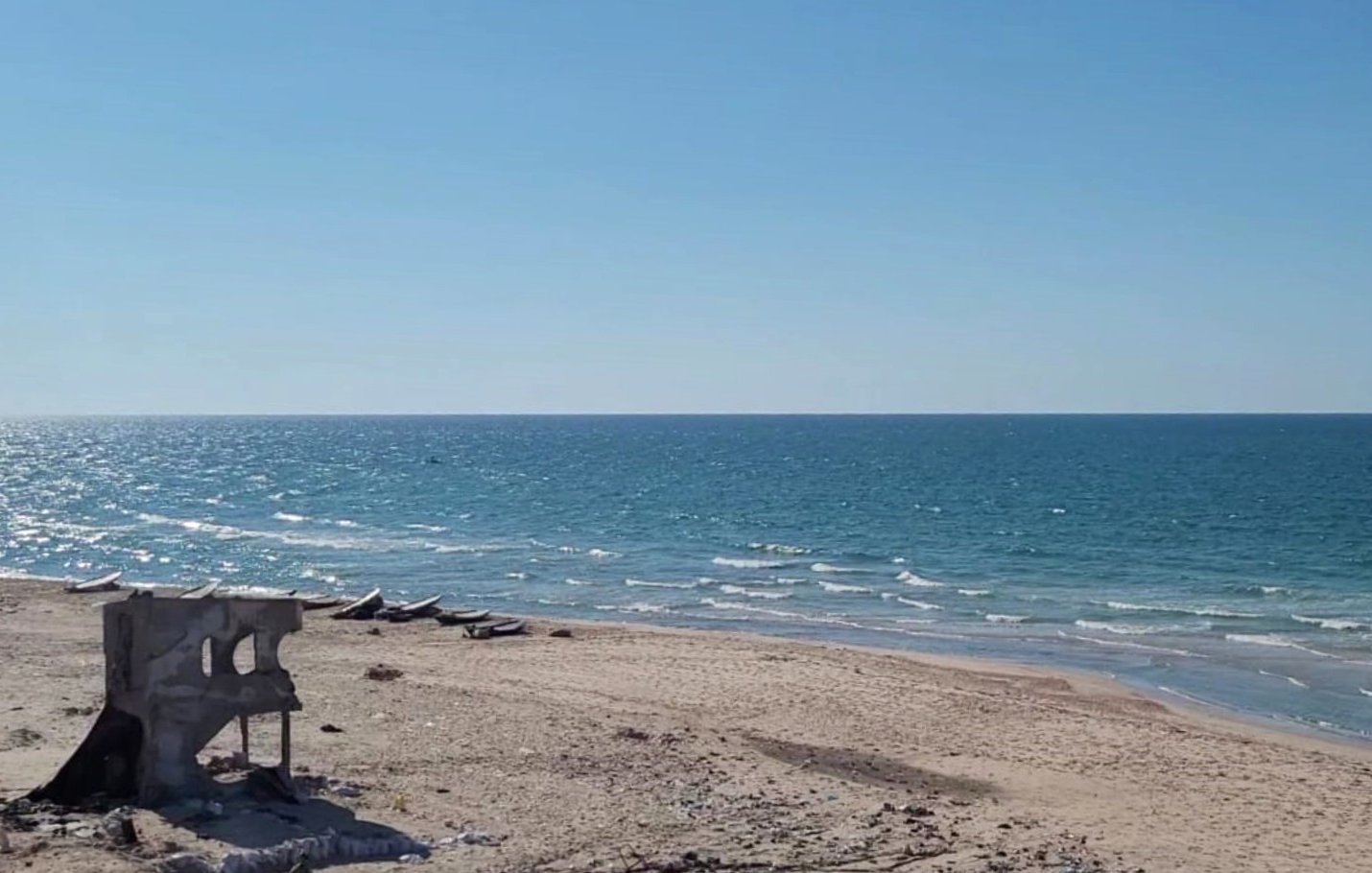 L'immagine mostra una spiaggia con sabbia chiara e un mare blu intenso sotto un cielo sereno e azzurro. In lontananza si possono vedere alcune imbarcazioni sulla superficie dell'acqua. Una struttura diroccata, grigia e in parte crollata, si trova sulla spiaggia, creando un contrasto con il paesaggio naturale circostante.