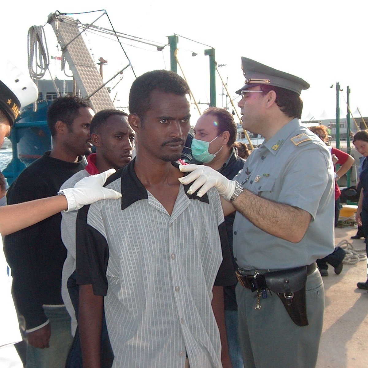 L'image montre une scène avec plusieurs personnes. Au premier plan, un homme se tient debout, apparemment en train d'être examiné par des agents portant des uniformes. Il y a un groupe d'autres personnes en arrière-plan, semblant également être supervisées. L'environnement suggère que cela se passe dans un port ou une zone maritime, avec des bateaux et des structures portuaires visibles. Les agents semblent s'occuper de la situation de manière attentive.