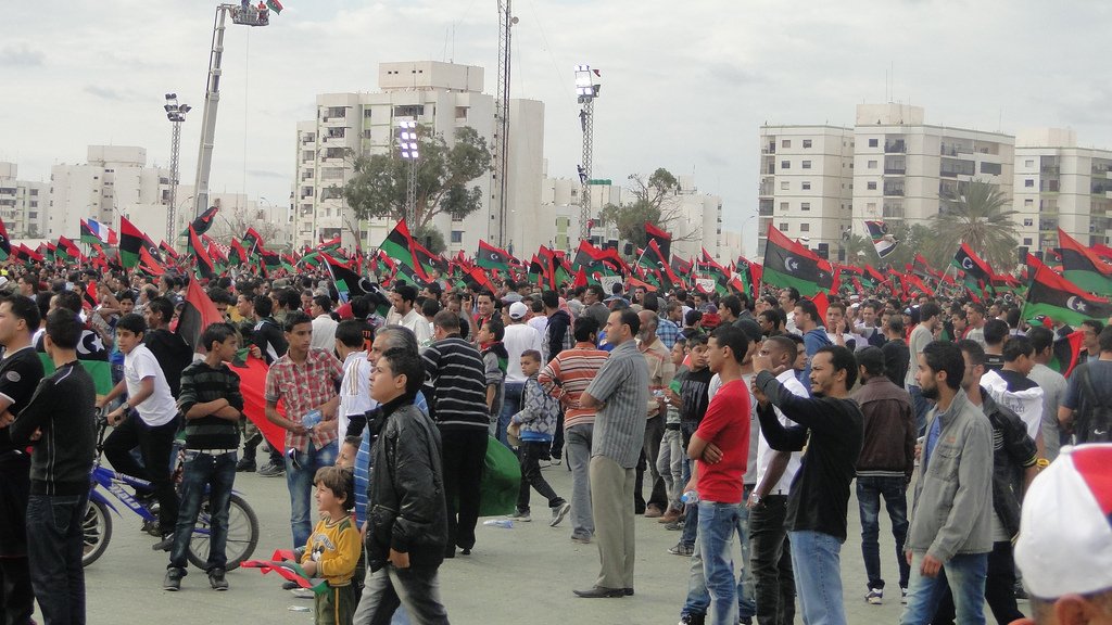 L'image montre une foule rassemblée dans un espace public, probablement lors d'un événement ou d'une célébration. De nombreuses personnes, dont des hommes, des femmes et des enfants, tiennent des drapeaux aux couleurs rouge, noir et vert, qui sont typiques du drapeau libyen. L'ambiance semble festive et dynamique, avec des bâtiments modernes en arrière-plan et des équipements techniques pour la sonorisation ou l'éclairage. On peut également voir des personnes sur des vélos, ajoutant une dimension de vie quotidienne à la scène.