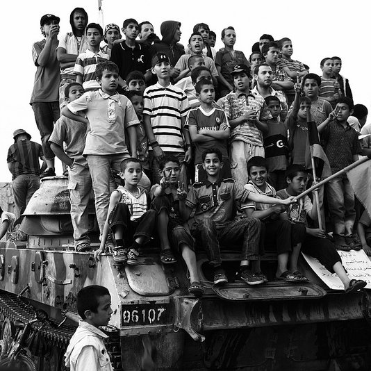 L'image montre un grand groupe d'enfants rassemblés sur un char, probablement dans un contexte de célébration ou de manifestation. Les enfants affichent diverses expressions, allant de la joie à la curiosité. Ils portent des vêtements colorés, souvent rayés, et certains tiennent un drapeau. En arrière-plan, on peut voir des bâtiments et des ruines, suggérant un environnement marqué par des conflits ou des événements historiques. L'image est en noir et blanc, ce qui accentue le côté dramatique de la scène.