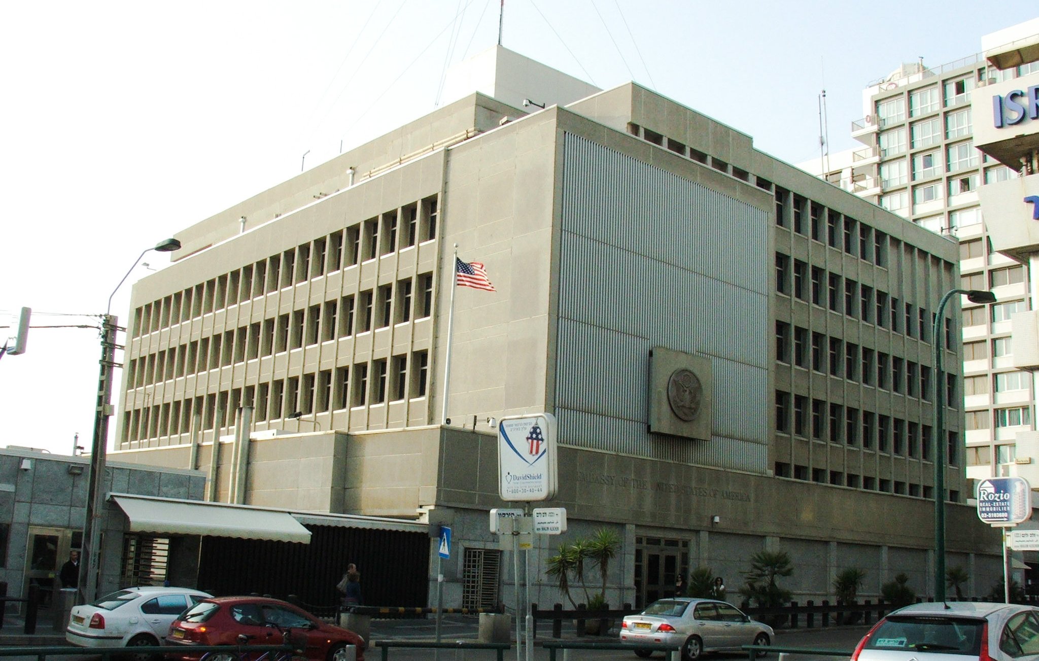 L'image montre un bâtiment moderne avec une architecture fonctionnelle. Il est en béton, avec plusieurs fenêtres rectangulaires sur la façade. Un drapeau américain est visible sur le toit, indiquant que c'est une représentation diplomatique. À l'avant du bâtiment, il y a des panneaux de signalisation et quelques véhicules garés le long de la route. L'environnement semble urbain, avec d'autres immeubles en arrière-plan.