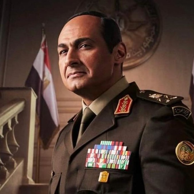 L'image montre un homme en uniforme militaire, vraisemblablement un officier supérieur. Il porte plusieurs décorations et insignes sur sa tenue, ce qui indique un haut rang. En arrière-plan, on aperçoit une architecture grandiose avec des drapeaux, ce qui suggère un environnement officiel, peut-être un gouvernement ou une institution militaire. L'expression de l'homme est sérieuse et déterminée.