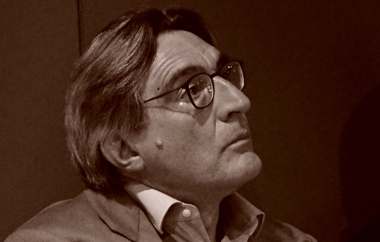 L'image montre un homme aux cheveux grisonnants, portant des lunettes et un costume clair. Il semble pensif, regardant quelque chose hors champ. L'éclairage et le ton de l'image lui donnent un aspect rétro, suggérant une ambiance contemplative.