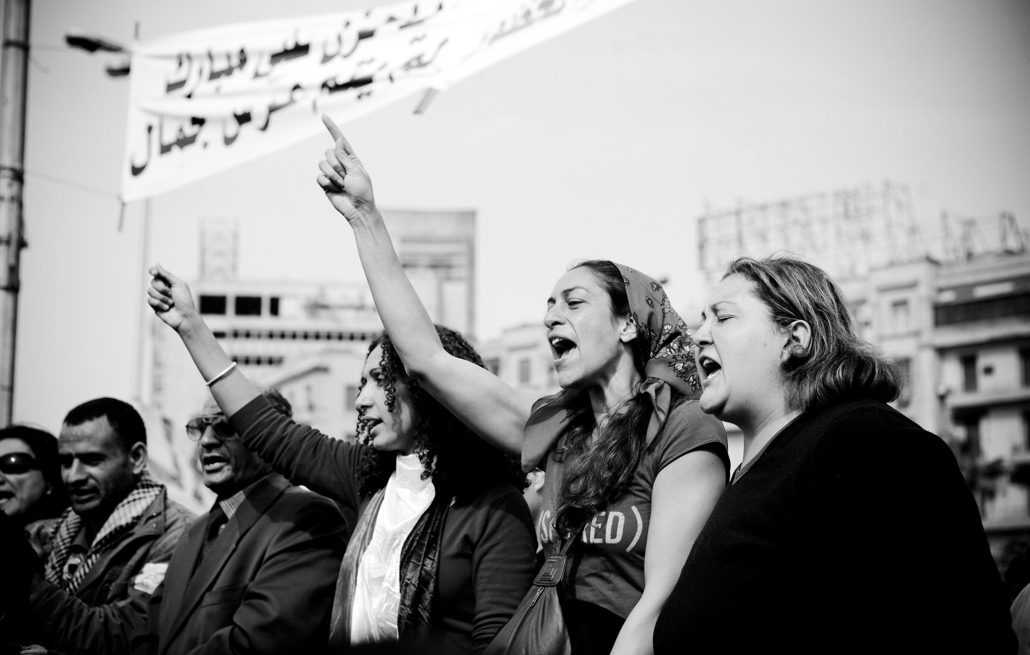L'image montre un groupe de personnes participant à une manifestation. On peut voir des femmes au premier plan levant le poing et chantant, exprimant des émotions fortes et de la détermination. L'ambiance semble dynamique et engageante. En arrière-plan, des banderoles apparaissent, suggérant un message ou une demande collective. La photo est en noir et blanc, ce qui accentue l'intensité et l'impact visuel de la scène.