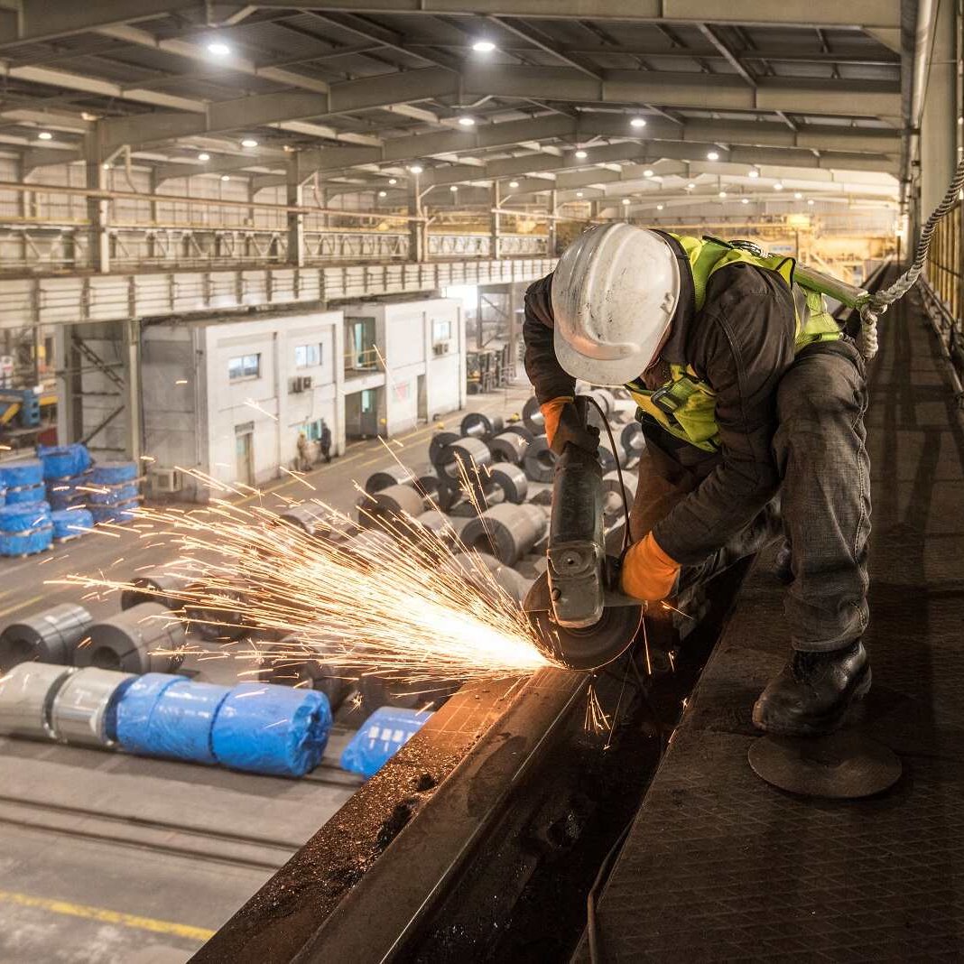 تظهر في الصورة عاملاً يرتدي خوذة وسماعات أذن، بينما يقوم بعملية قطع أو طحن المعادن في مصنع. تتطاير الشرر أثناء عملية العمل، مما يضيف إلى الحركة والديناميكية في المكان. خلف العامل، يُمكن رؤية مساحة واسعة من المصنع تضم معدات وعجلات مغطاة بأقمشة زرقاء، مما يشير إلى بيئة صناعية نشطة. الإضاءة في الصورة جيدة، مما يبرز تفاصيل العمل والبيئة المحيطة.