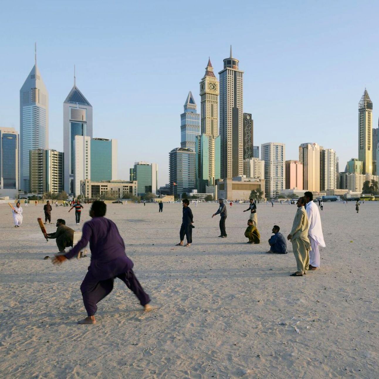 الصورة تُظهر مشهدًا حضريًا لمدينة حديثة، حيث تبرز ناطحات السحاب والمباني الشاهقة. في المقدمة، يوجد مجموعة من الأشخاص يمارسون أنشطة مختلفة على رمال منطقة مفتوحة، ما يوحي بأجواء حيوية. الخلفية تُظهر المدينة مع سماء زرقاء تتخللها أشعة الشمس، مما يضفي لمسة جميلة على المشهد.