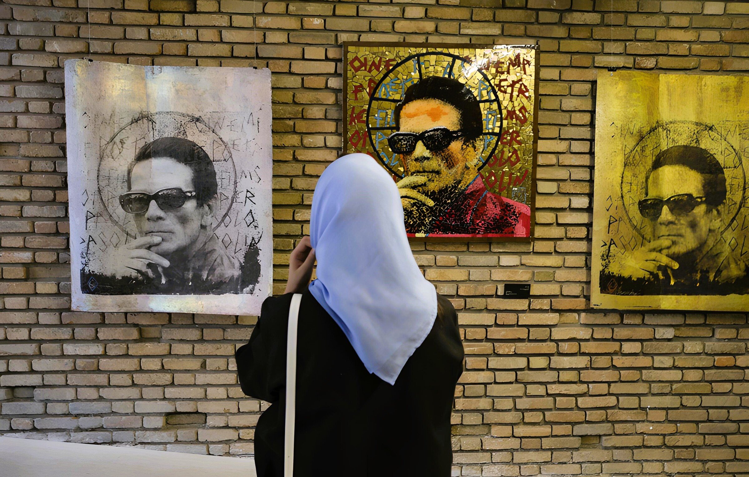 L'image montre une femme portant un hijab en train d'observer des œuvres d'art accrochées à un mur en briques. Les tableaux représentent un homme avec des lunettes de soleil, entouré de motifs et de couleurs variés. L'ambiance semble artistique et contemporaine, avec un jeu de lumière qui attire l'œil vers les œuvres.