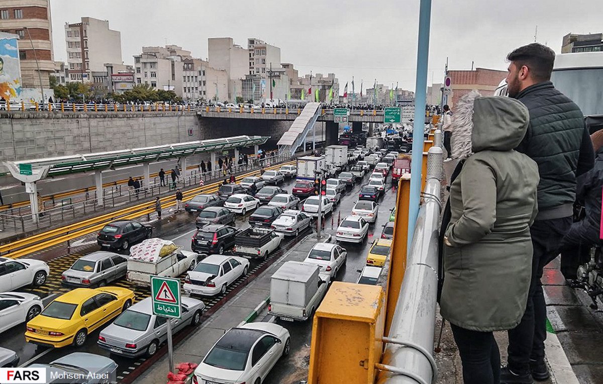 تصویر نشان‌دهنده یک صحنه شلوغ از ترافیک در یک خیابان شهری است. در سمت چپ تصویر، افرادی در حال تماشا از بالا ایستاده‌اند. در پایین، خطوط خودروها به وضوح قابل مشاهده است که نشان‌دهنده تجمع و ترافیک سنگین است. بقیه عناصر شهری مانند ساختمان‌ها و علائم راهنمایی نیز در پس‌زمینه دیده می‌شوند. هوای ابری و احتمالا بارانی به دید فضا و حس و حال تصویر اضافه می‌کند.