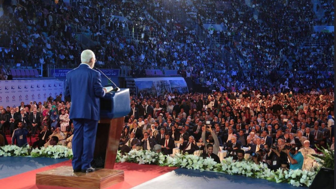 L'image montre un homme debout sur une scène, s'adressant à une foule nombreuse. Il est en costume et se tient derrière un pupitre, probablement en train de donner un discours. La salle est pleine, avec des centaines de personnes assises en face de lui, certaines regardant attentivement. L'éclairage crée une ambiance dramatique, mettant en valeur à la fois le conférencier et le public. Des décorations florales sont visibles au premier plan, ajoutant une touche d'élégance à l'événement.