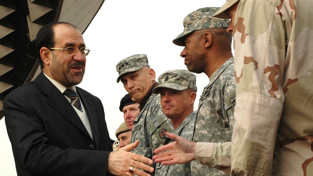 L'image montre un homme en costume qui serre la main de plusieurs militaires. L'homme semble être en discussion amicale avec eux. Les soldats portent des uniformes militaires, et l'environnement semble être une zone aéroportuaire, avec un avion visible en arrière-plan. Les expressions des militaires sont sérieuses, tandis que l'homme en costume sourit. Ce moment pourrait symboliser une collaboration ou une rencontre officielle.