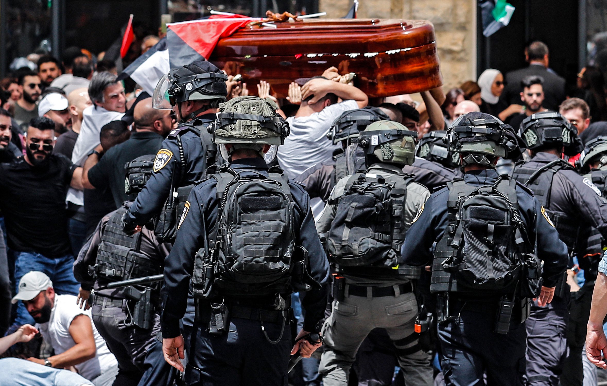 تظهر الصورة مشهدًا من حدث عام حيث يحدث تداخل بين مجموعة من الناس وقوات الأمن. يتم حمل تابوت خشبي من قبل المشاركين في الهتافات، بينما يحيط بهم رجال شرطة يرتدون تجهيزات خاصة. الخلفية تضم أعلامًا، مما يشير إلى أن هذه الفعالية قد تكون مرتبطة بمسألة وطنية أو سياسية. الجو يبدو مشحونًا بالانفعالات، مع وجود حشد من الناس في حالة من التوتر.