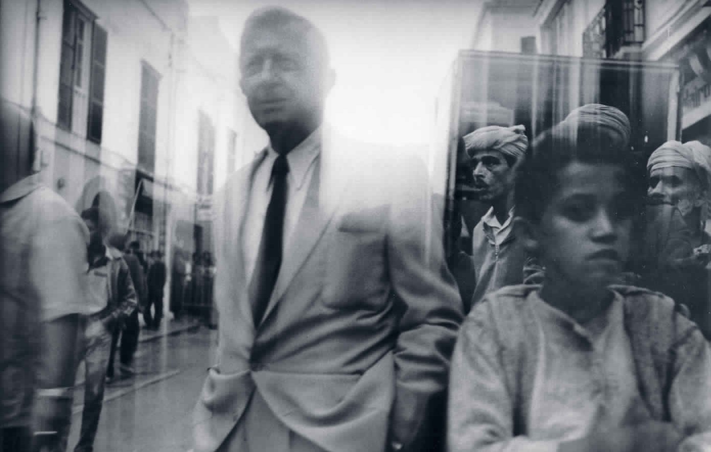 L'image semble capturer une scène de rue en noir et blanc, où se mêlent des figures humaines. Au premier plan, un homme en costume clair se tient debout, semblant légèrement détaché du reste de la foule. En arrière-plan, on aperçoit plusieurs personnes, probablement des habitants, qui regardent vers la caméra. L'ambiance est floue, ce qui donne une impression de mouvement et de vie dynamique dans la rue. Les vêtements traditionnels des personnes en arrière-plan contrastent avec le style moderne de l'homme au premier plan, créant une image intéressante et expressive.