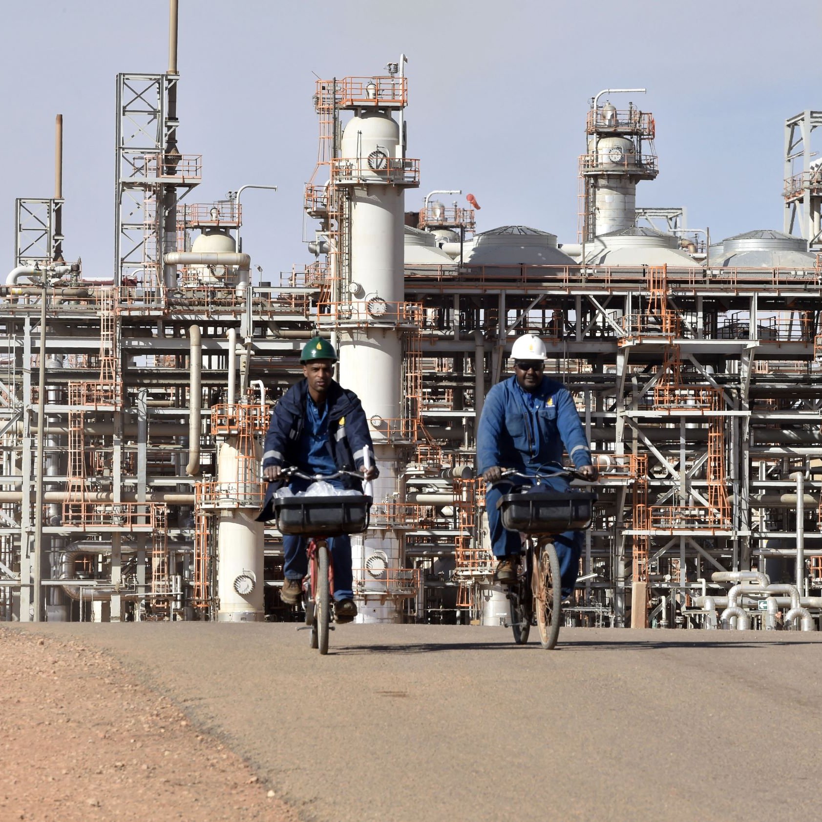 L'image montre deux hommes à vélo, portant des vêtements de travail et des casques, qui circulent sur une route. En arrière-plan, on peut voir une installation industrielle avec de grandes structures métalliques et des pipelines, typiques d'une raffinerie ou d'un complexe pétrochimique. Le ciel est clair et les installations semblent actives, témoignant d'un environnement de travail industriel.