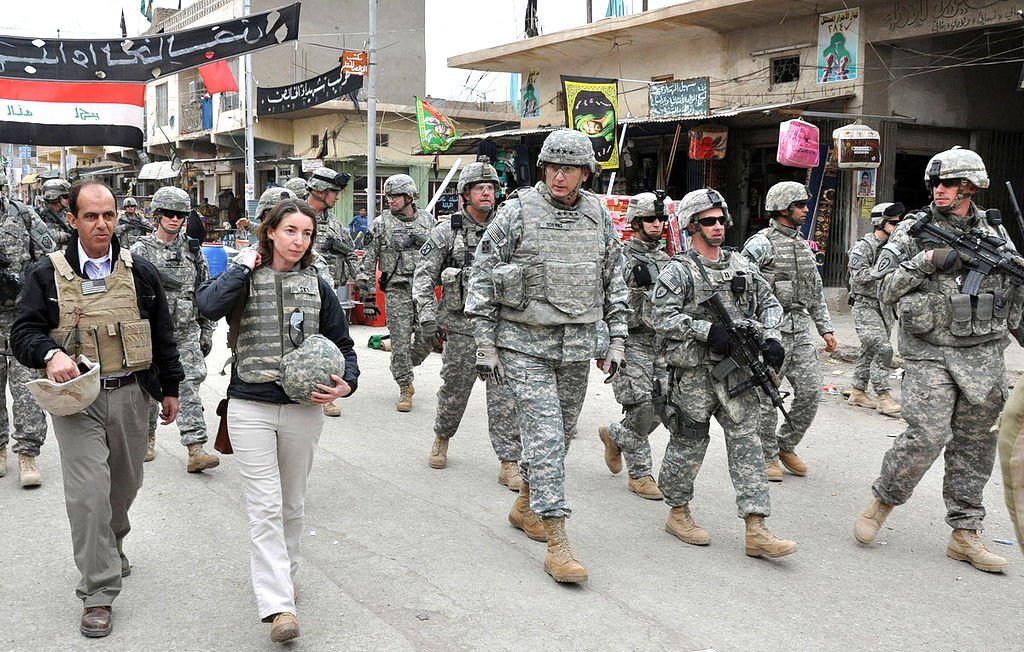L'image montre un groupe de soldats en uniforme militaire marchant dans une rue. Ils semblent être dans une zone urbaine, possiblement dans un pays en conflit. Au premier plan, on voit une femme en vêtements civils, portant un gilet pare-balles, marchant à côté d'un homme également en tenue civile. Le décor autour d'eux comprend des bâtiments, des panneaux et d'autres signes de vie urbaine. L'atmosphère semble sérieuse, reflétant un environnement potentiellement tendu.