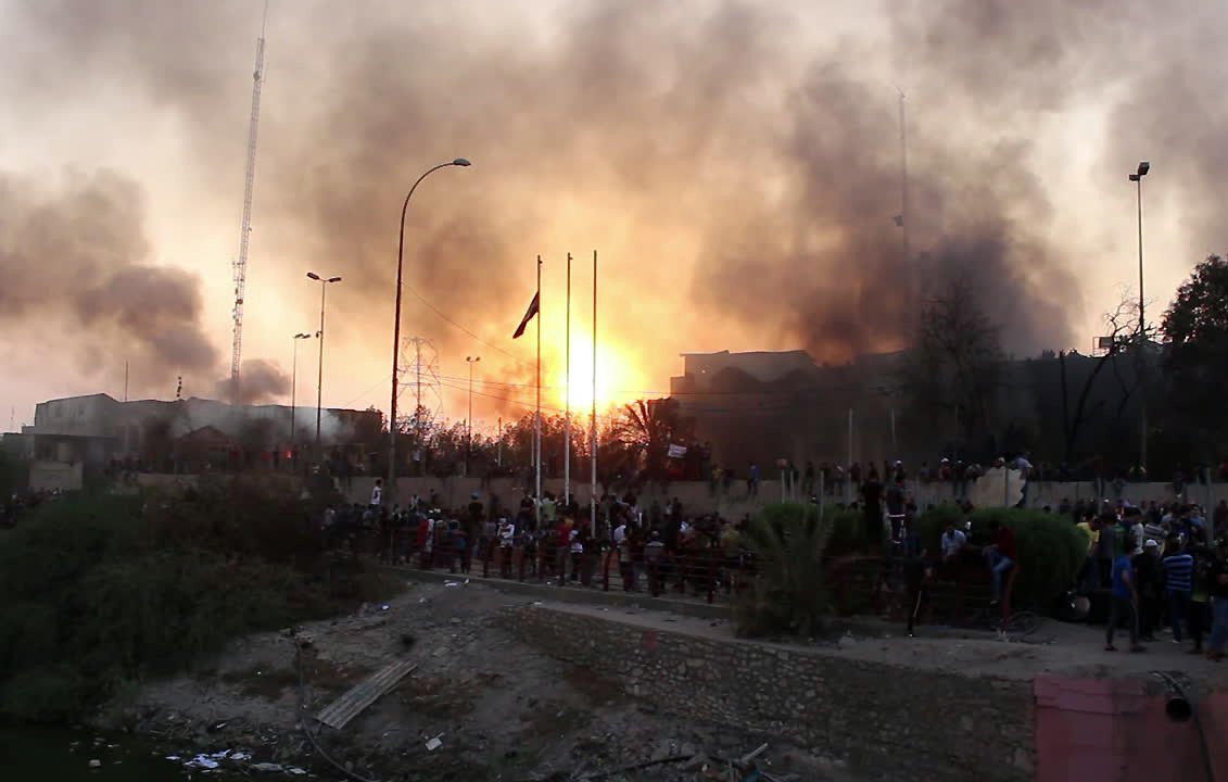 L'image montre une scène tumultueuse, probablement au cours d'un événement de protestation ou de conflit. On peut voir de la fumée s'élever dans le ciel, créant une atmosphère chaotique. À l'arrière-plan, un grand incendie semble se produire, projetant une lumière orange intense. Au premier plan, un groupe de personnes se rassemble, probablement en train d'observer ou de participer à l'événement. La présence de poteaux et de drapeaux suggère un espace urbain, peut-être une place publique ou un bord de fleuve. L'ensemble donne une impression d'urgence et de tension.