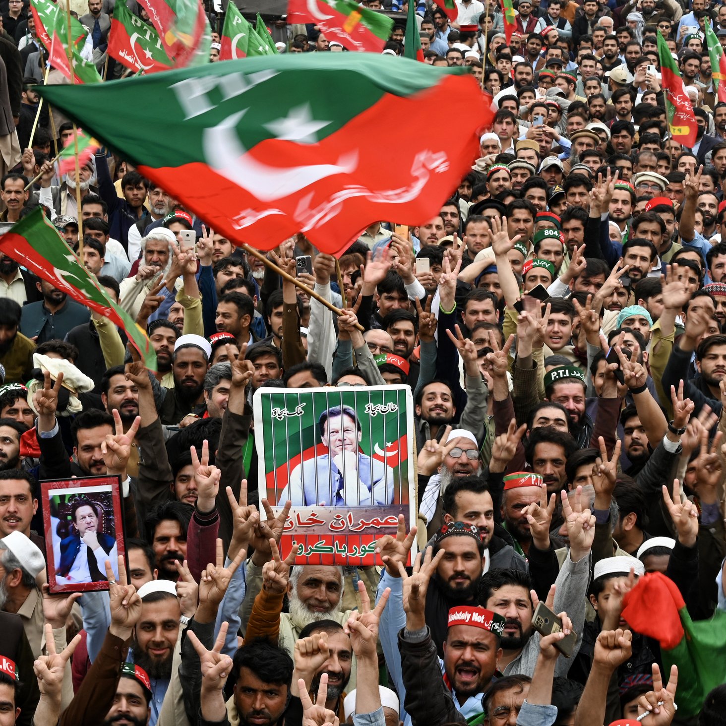L'image montre une grande foule rassemblée lors d'un rassemblement. De nombreuses personnes brandissent des drapeaux, en particulier le drapeau du Parti Pakistan Tehreek-e-Insaf (PTI). On peut voir des manifestants levant les bras avec des gestes de victoire, ainsi que des pancartes et des portraits de leaders politiques. L'ambiance semble dynamique et engageante, avec des cris et des slogans de la part des partisans.