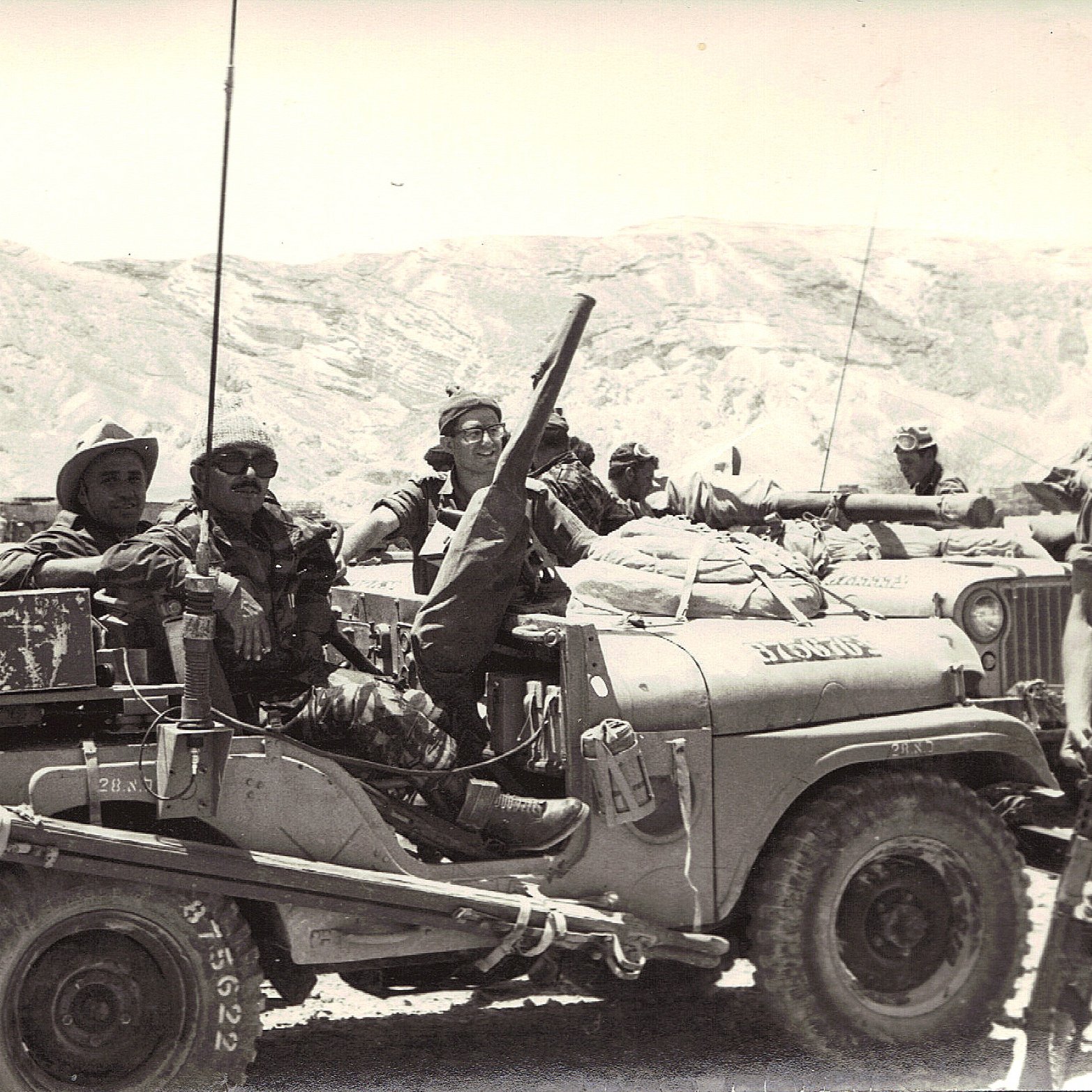 L'image montre un groupe de soldats réunis autour d'un véhicule militaire, probablement une jeep. Ils semblent engagés dans une mission, avec des équipements militaires visibles sur le véhicule. Le paysage en arrière-plan est aride, avec des collines ou des montagnes. Les soldats prennent la pose, certains souriant, et semblent confiants dans leur environnement. L'atmosphère dégage un sentiment de camaraderie et de détermination.