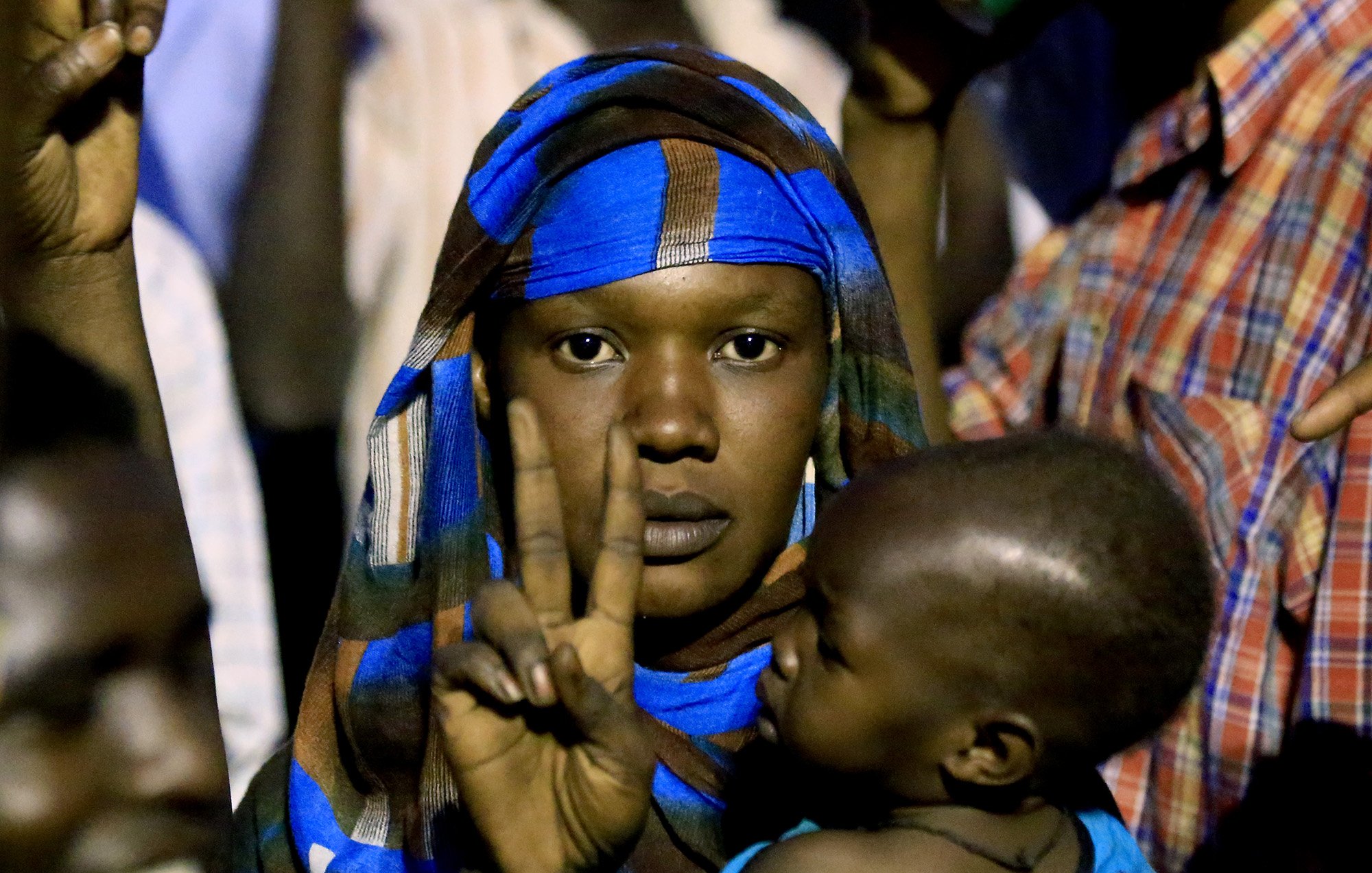 در این تصویر، زنی با یک شال رنگارنگ و نقوش آبی در حال نشان دادن علامت پیروزی (دو انگشت) است. او یک کودک را در آغوش دارد که به سمت او نگاه می‌کند. پس‌زمینه تصویر شامل افرادی است که در حال تماشا هستند و نور محیط کمی کم است. حالت زن نشان‌دهنده اعتماد به نفس و امیدواری است.