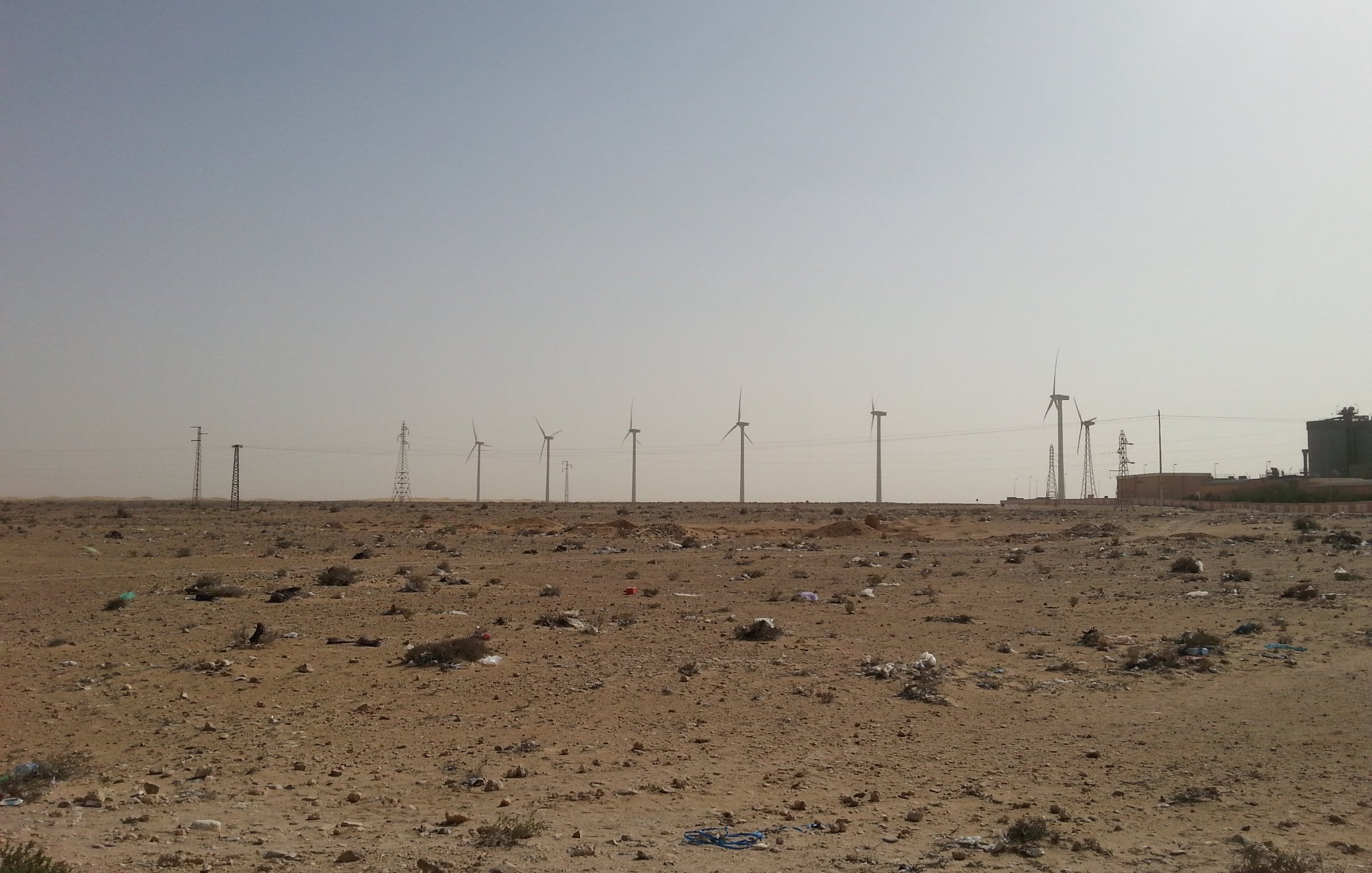 L'image montre un paysage désertique, caractérisé par un terrain aride avec des débris, probablement des déchets, éparpillés sur le sol. À l'arrière-plan, on peut voir plusieurs éoliennes qui s'élèvent vers le ciel clair. Des pylônes électriques sont également visibles, ajoutant une touche industrielle à ce paysage naturel. Le ciel est dégagé, ce qui accentue l'effet désertique de la scène.