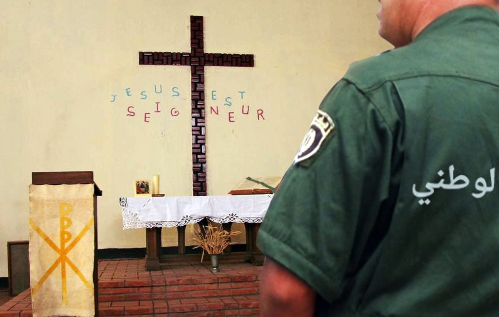 تظهر الصورة مشهداً داخل كنيسة، حيث يتواجد صليب خشبي كبير على الحائط. خلف الصليب، توجد كلمات مكتوبة باللغة الفرنسية تقول "يسوع هو الرب" (Jésus est Seigneur). في الجهة اليمنى من الصورة، يقف رجل يرتدي زيًا رسميًا ويرتدي شعارًا يدل على "الدرك الوطني". يبدو أن الرجل يراقب المكان حوله. الأجواء تعكس مزيجًا من الروحانية والتواجد الأمني.
