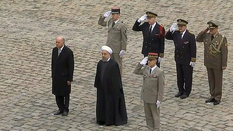 الصورة تظهر مجموعة من الأشخاص في موقف رسمي. يوجد شخصان في المقدمة، أحدهما في بدلة رسمية والآخر يرتدي الزي التقليدي. خلفهم يوجد عدد من الضباط العسكريين بزيهم الرسمي، يقومون بتحية عسكرية. الأرضية مكونة من حجارة مرصوفة. الجو يبدو رسميًا وجادًا، مما يشير إلى حدث ذو أهمية سياسية أو عسكرية.