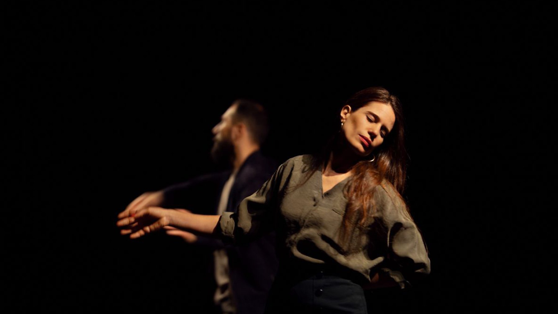 Deux danseurs se déplacent sur scène dans une lumière tamisée. Une femme, les yeux fermés et une expression de sérénité, se tient légèrement en avant, tandis qu'un homme, derrière elle, semble effectuer un mouvement fluide avec ses bras. L'atmosphère est intimiste et expressive.