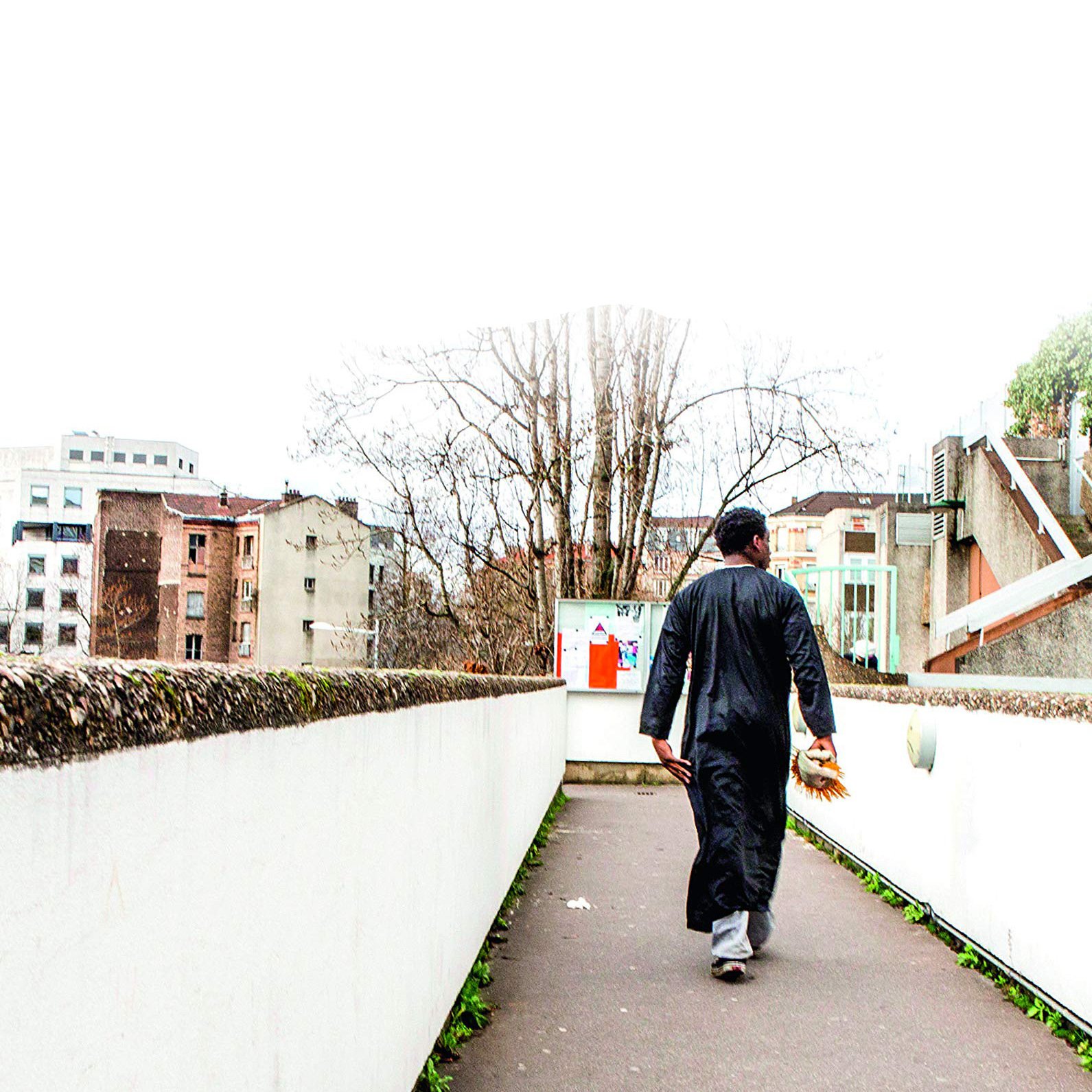 L'image montre un homme marchant sur un chemin urbain. Il porte une tenue traditionnelle, une sorte de longue robe noire. En arrière-plan, on peut voir des bâtiments modernes et anciens, ainsi que des arbres dépouillés. L'environnement semble calme, avec une lumière diffuse, peut-être un jour nuageux. L'homme avance vers l'horizon, de dos, ce qui donne une impression de solitude et de contemplation.